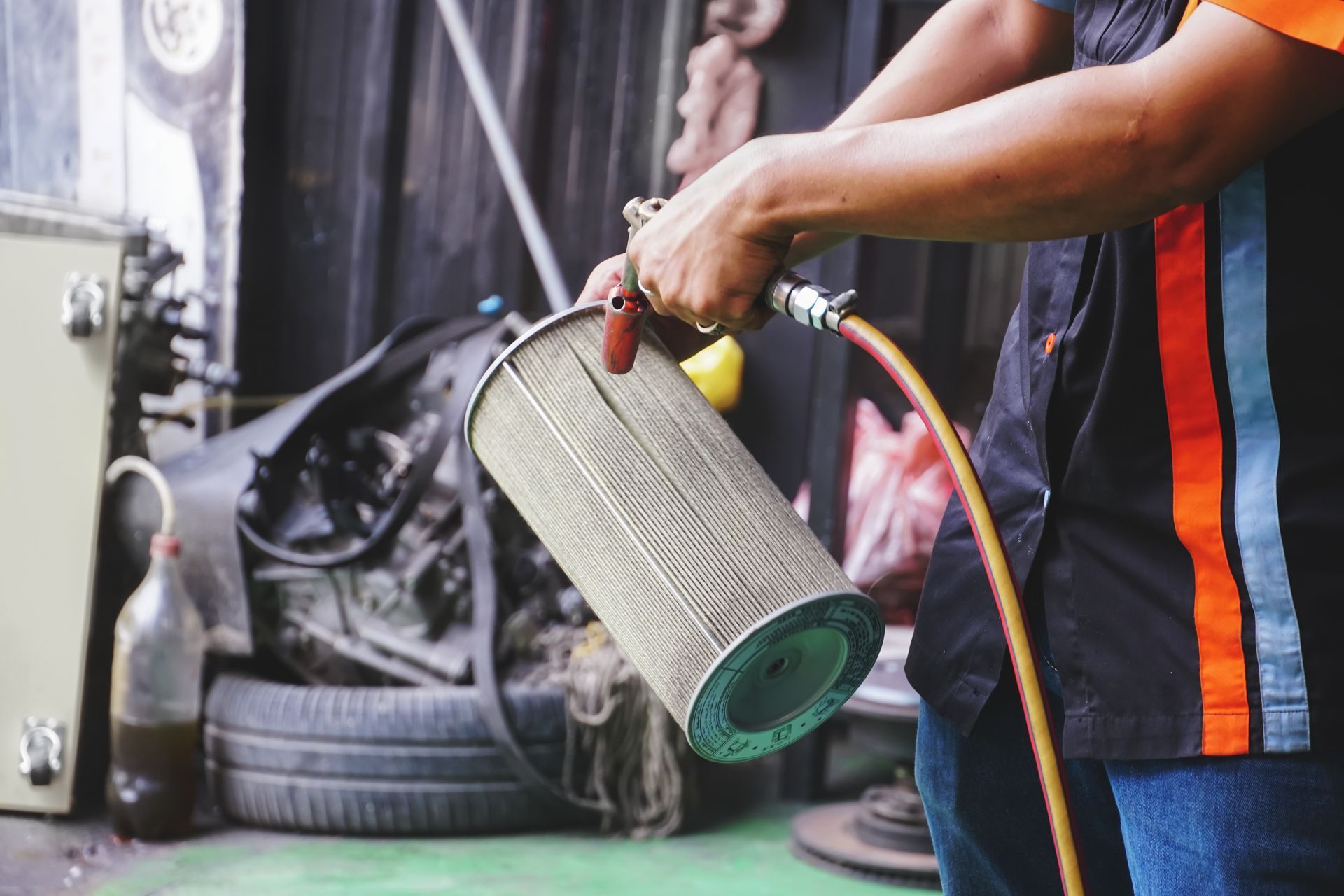 Une personne tient un grand cylindre filtrant près d'un pneu de voiture dans un garage, avec un tuyau d'aspirateur fixé.
