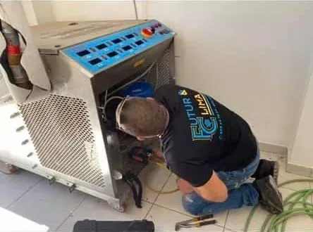 Un técnico se arrodilla junto a una gran máquina azul y blanca, inspeccionando el cableado en un cuarto de servicio.