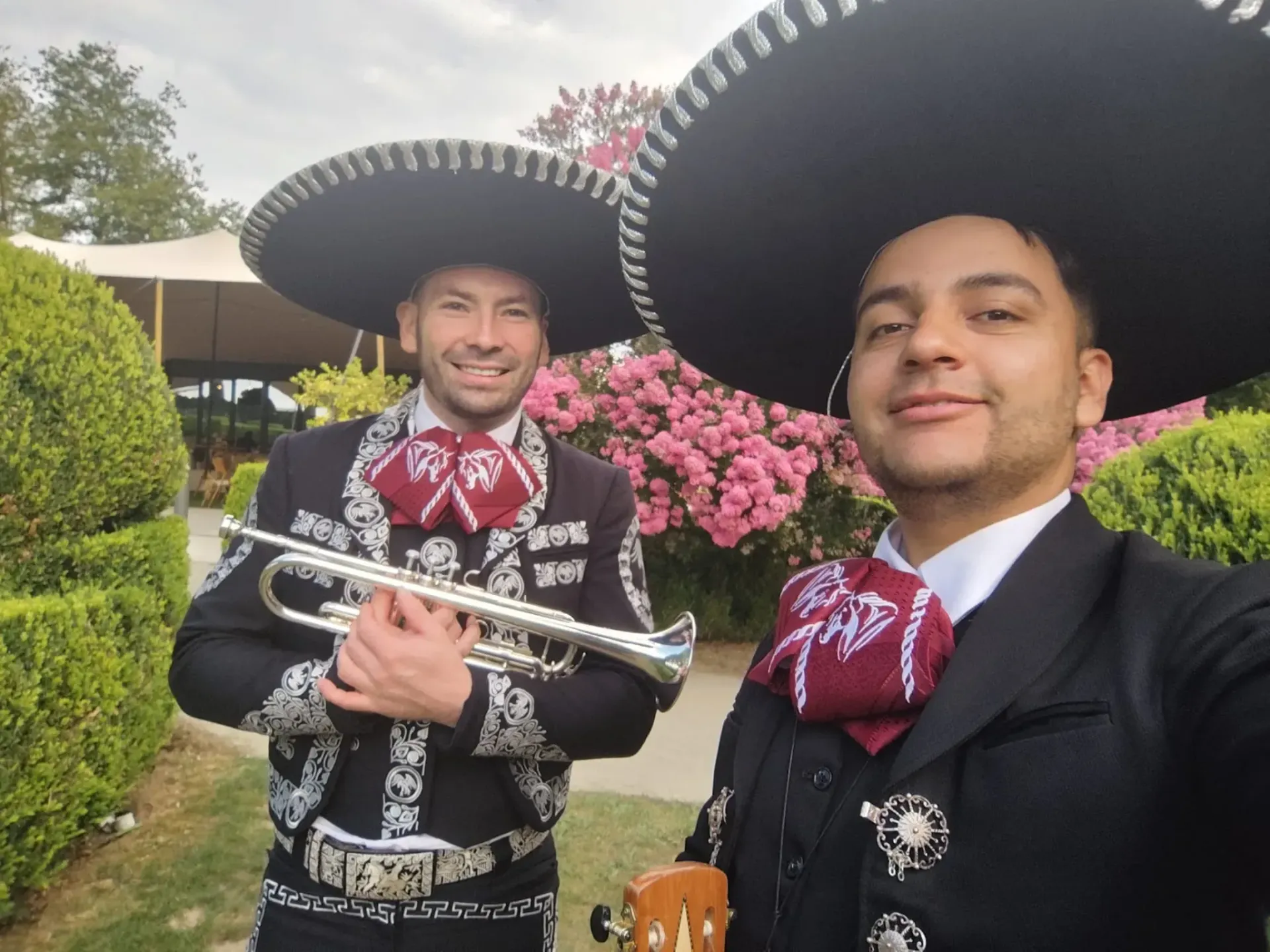 Dos hombres vestidos de mariachi posan al aire libre, uno de ellos sosteniendo una trompeta, cerca de arbustos y una tienda de campaña.