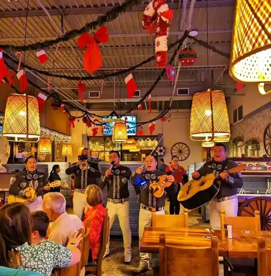 Un mariachi toca en un restaurante decorado con motivos festivos. La gente observa y cena.