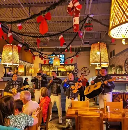 Un mariachi toca en un restaurante decorado con motivos festivos. La gente observa y cena.