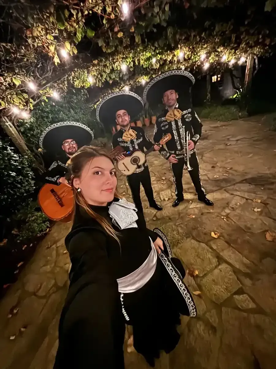 Una mujer se toma una selfie con un mariachi bajo luces de colores. Los músicos visten trajes tradicionales.