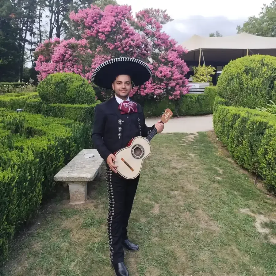 Músico mariachi con atuendo tradicional, sosteniendo una guitarra, de pie en un jardín con un fondo floral.