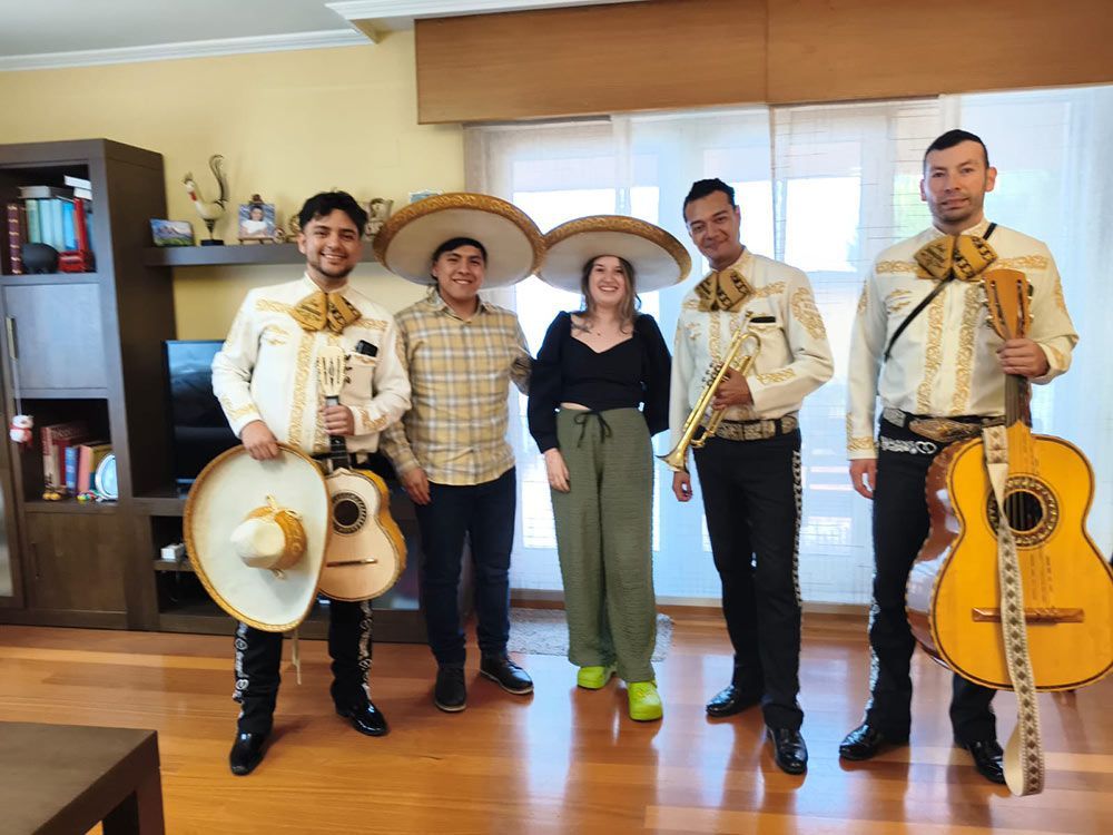 Cinco personas vestidas con trajes tradicionales de charro mexicano posando en el interior, con guitarras y sombrero, en una habitación luminosa.