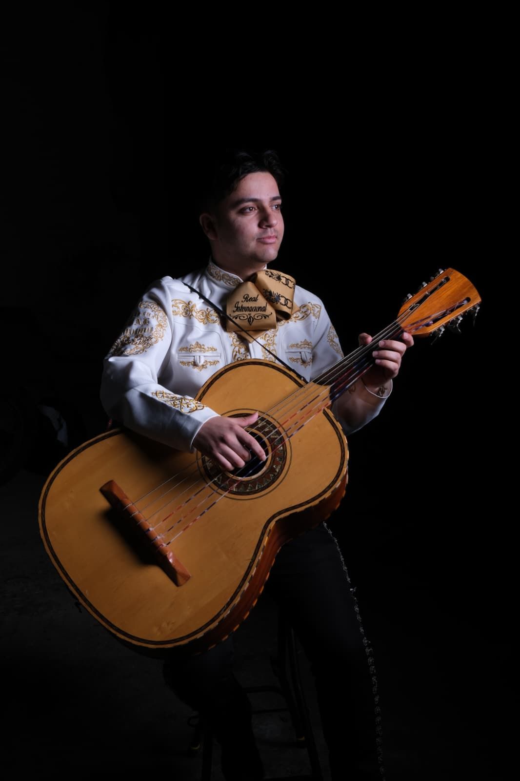 Músico con camisa blanca bordada sosteniendo una guitarra sobre un fondo negro.