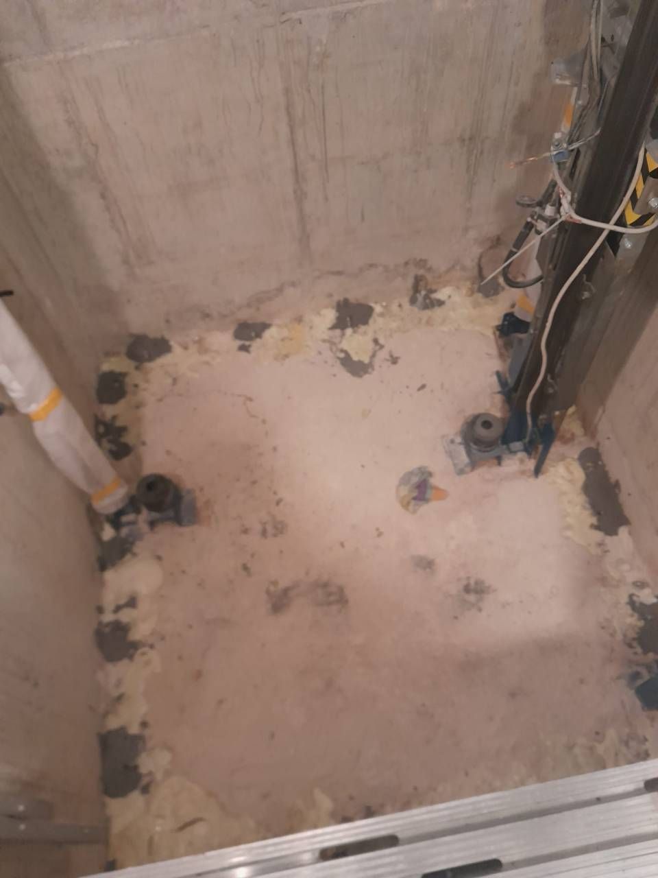 Pièce brute aux murs de béton, avec de la mousse isolante sur les bords du sol. On aperçoit divers tuyaux et câbles électriques.