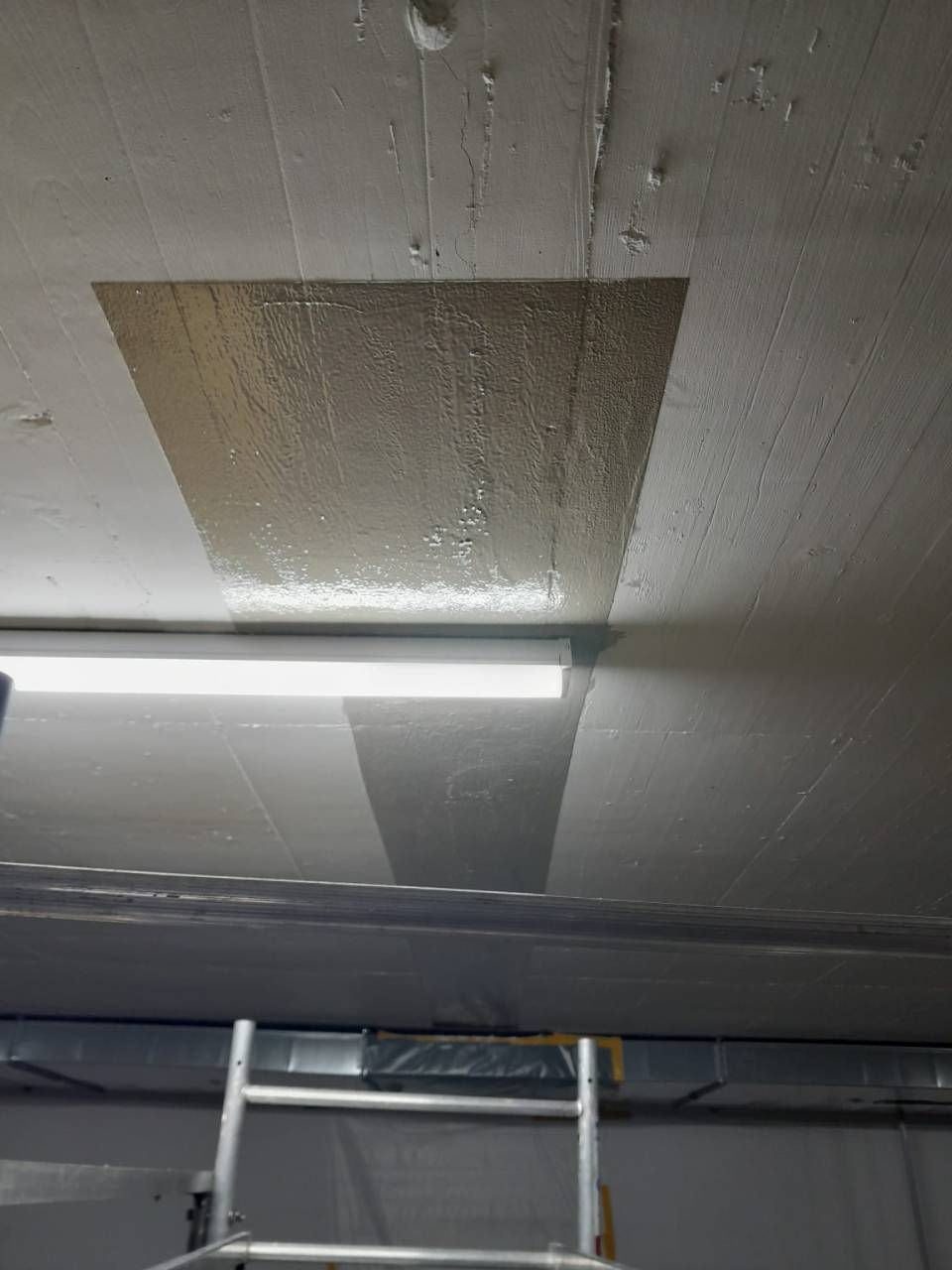 Plafond en béton gris avec une réparation rectangulaire, éclairé par une lampe fluorescente et une échelle en dessous.
