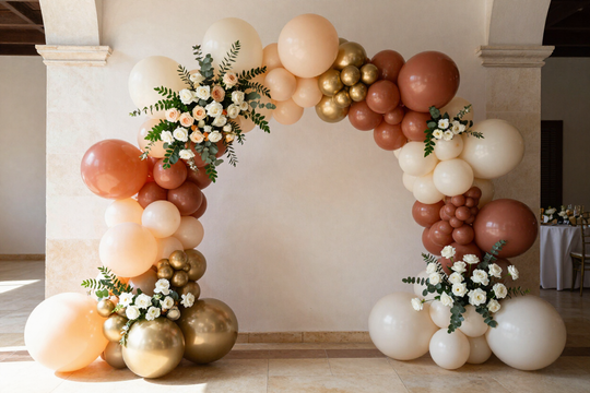 Arco de globos en tonos melocotón, blanco y dorado, con detalles florales, enmarcando una pared vacía en una habitación