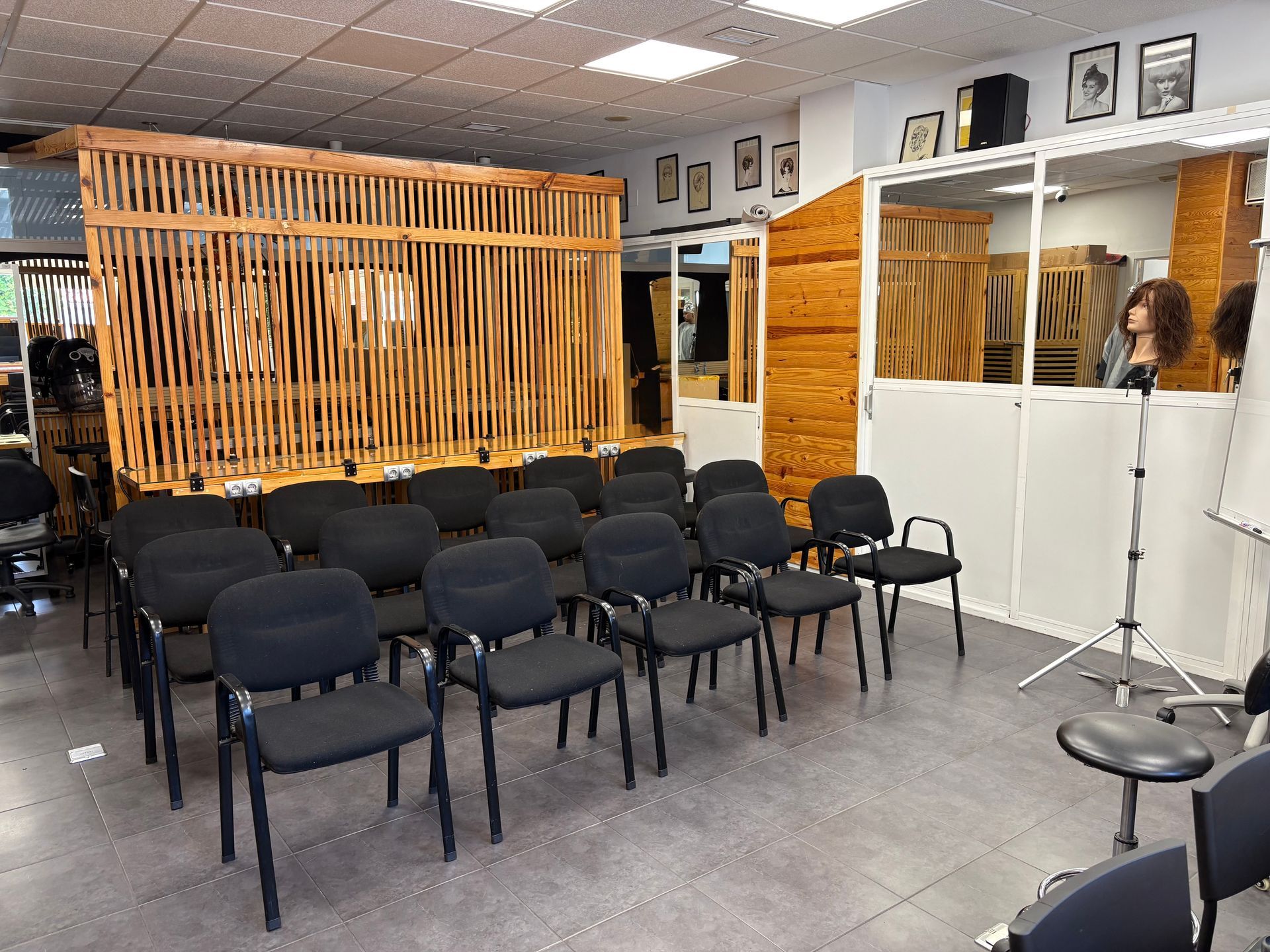 Aula vacía con filas de sillas negras, paredes blancas, separador de listones de madera y un soporte para micrófono a la derecha.