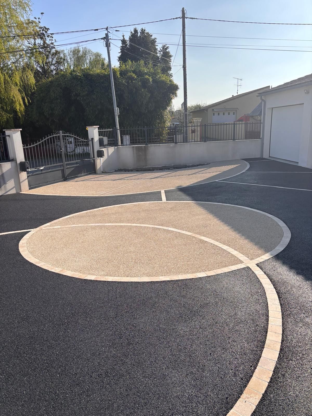 Allée asphaltée avec design circulaire beige, clôture, garage, arbres et lignes électriques.