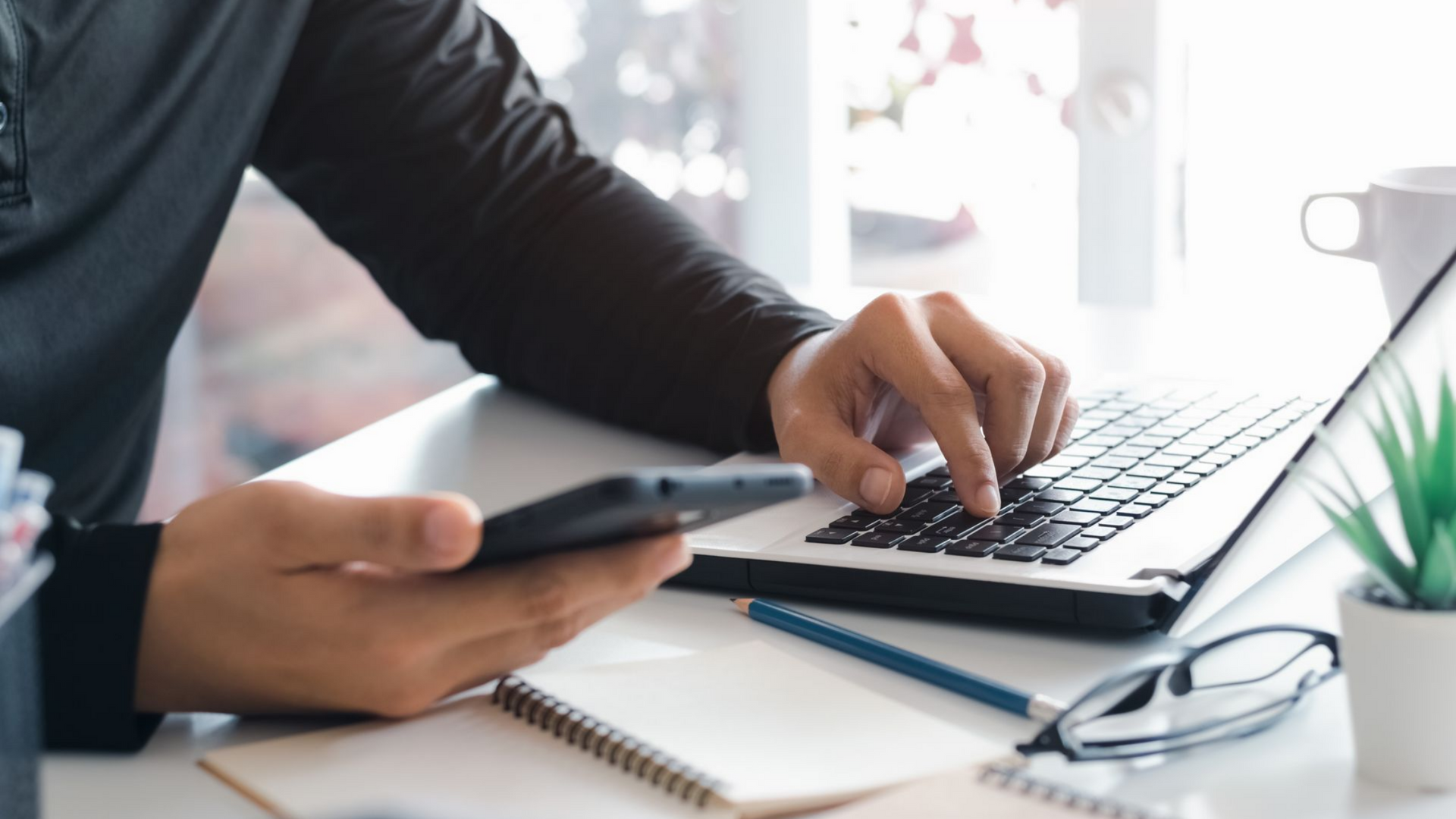 Une personne utilise un ordinateur portable et un smartphone à un bureau. Ses mains tapent sur le clavier, tandis que l'autre tient son téléphone. Sur une surface blanche, on aperçoit une tasse de café, une plante et des lunettes.