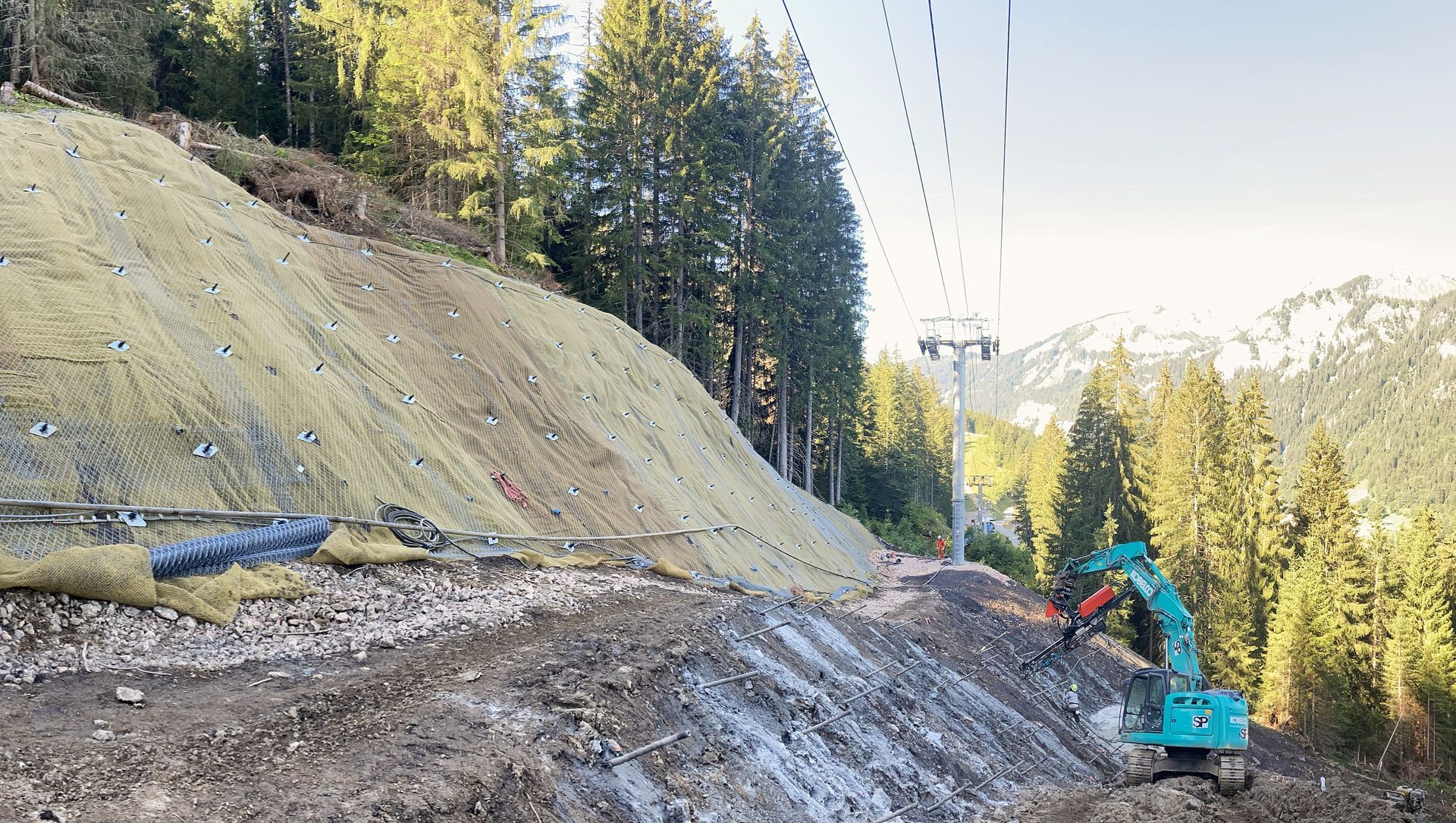 Chantier de construction à flanc de montagne, avec une excavatrice, des filets anti-érosion et un pylône de téléski.