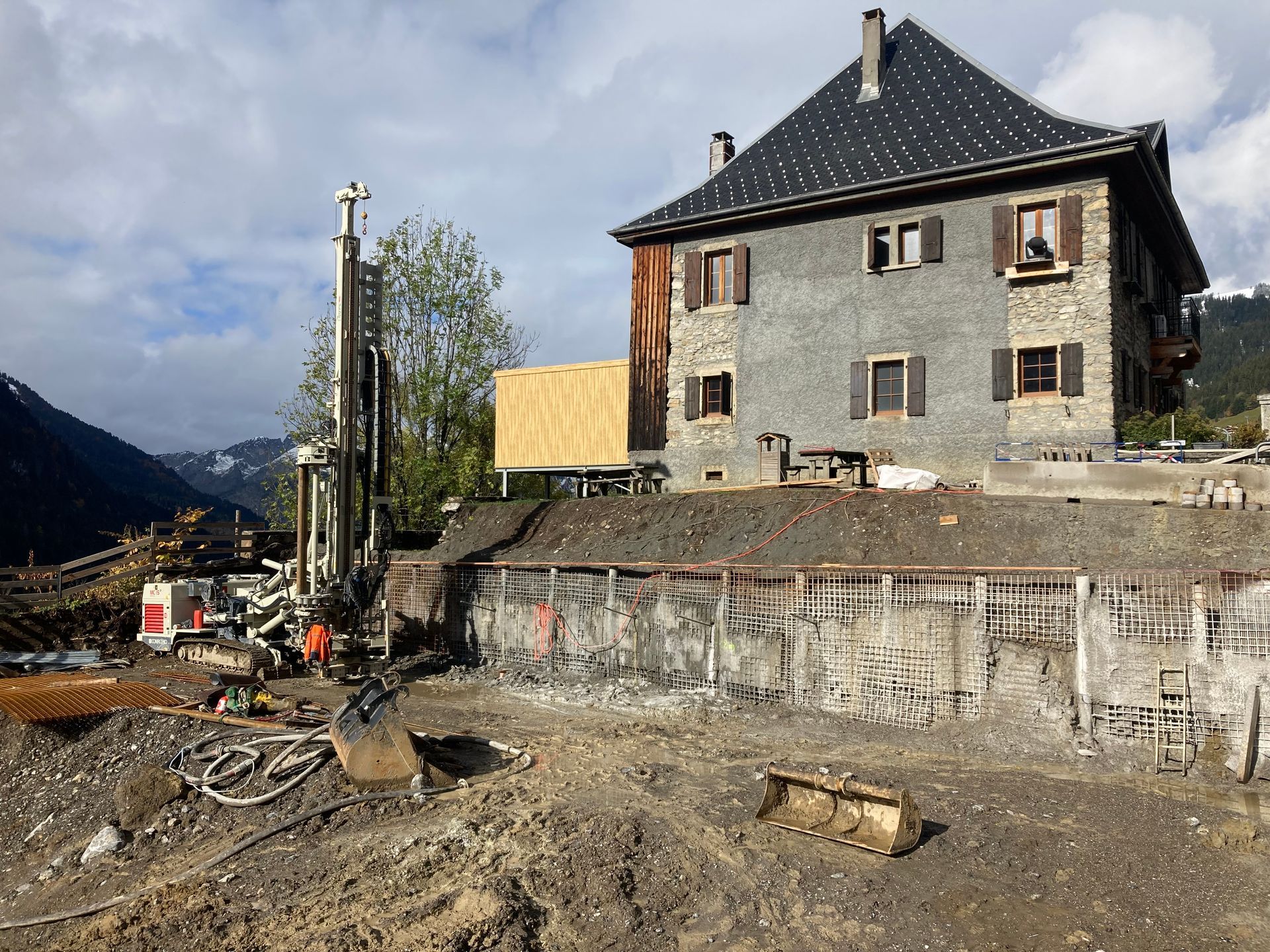 Chantier de construction avec matériel de forage près d'un immeuble de plusieurs étages et d'une montagne en arrière-plan.