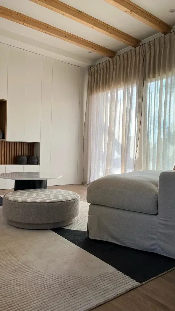 Una sala de estar con sofá, otomana, mesa de café y grandes ventanales.