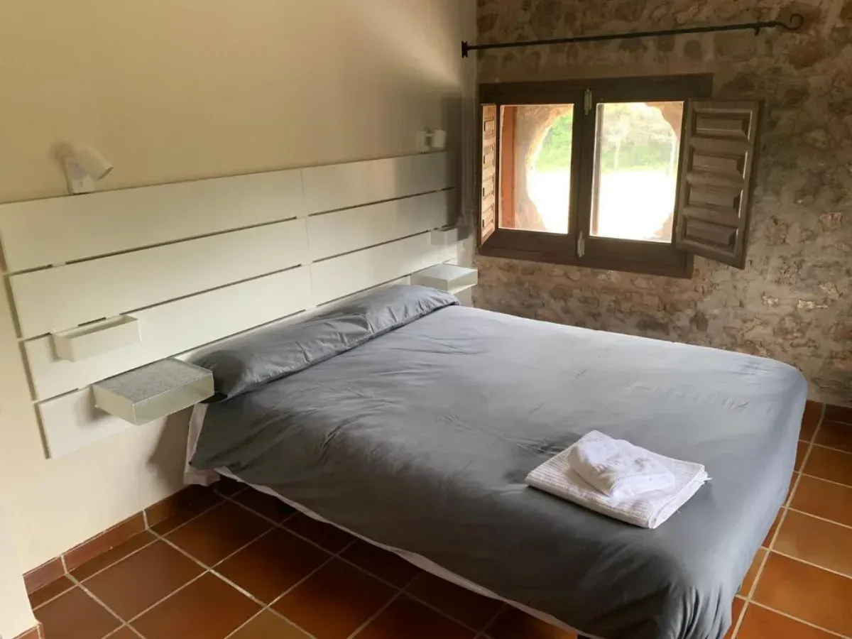 Habitación pequeña con cama gris, estantes blancos, pared de piedra y ventana con contraventanas.