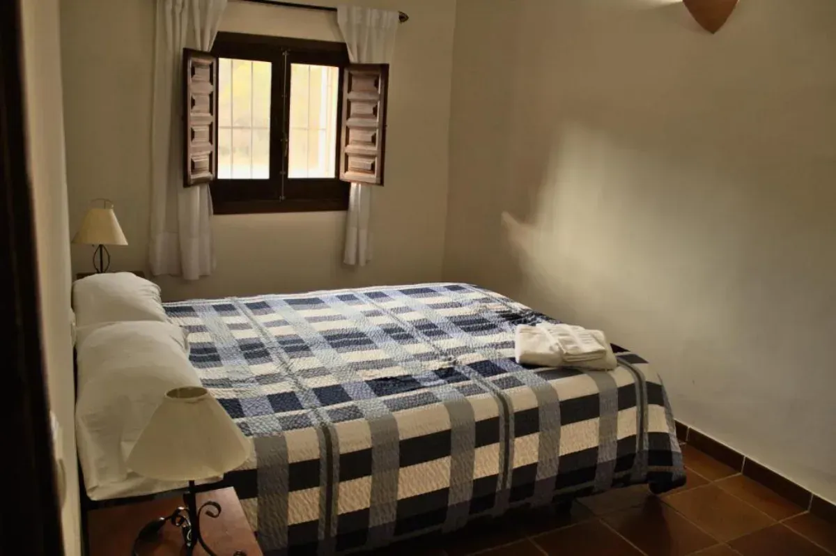 Habitación pequeña con cama individual, manta a cuadros azules, ventana abierta y paredes beige.