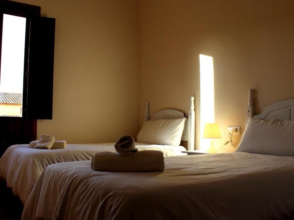 Habitación soleada con dos camas individuales, paredes de color beige y una pequeña ventana a la izquierda.