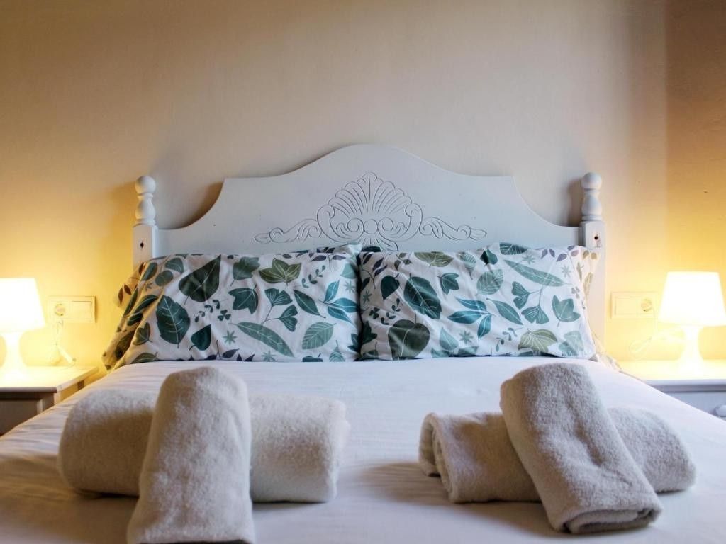 Cama blanca con cojines estampados con flores, toallas enrolladas y lámparas de noche en un dormitorio con iluminación tenue.