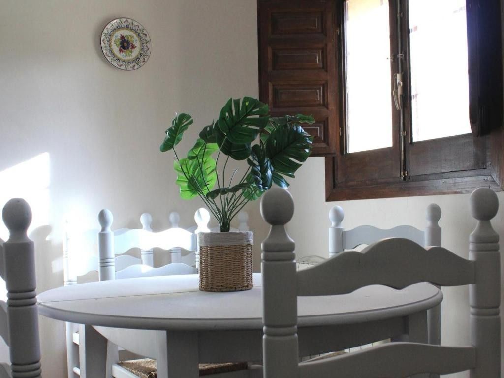 Mesa redonda blanca con sillas en una habitación luminosa, junto a una ventana y un centro de mesa con una planta frondosa.
