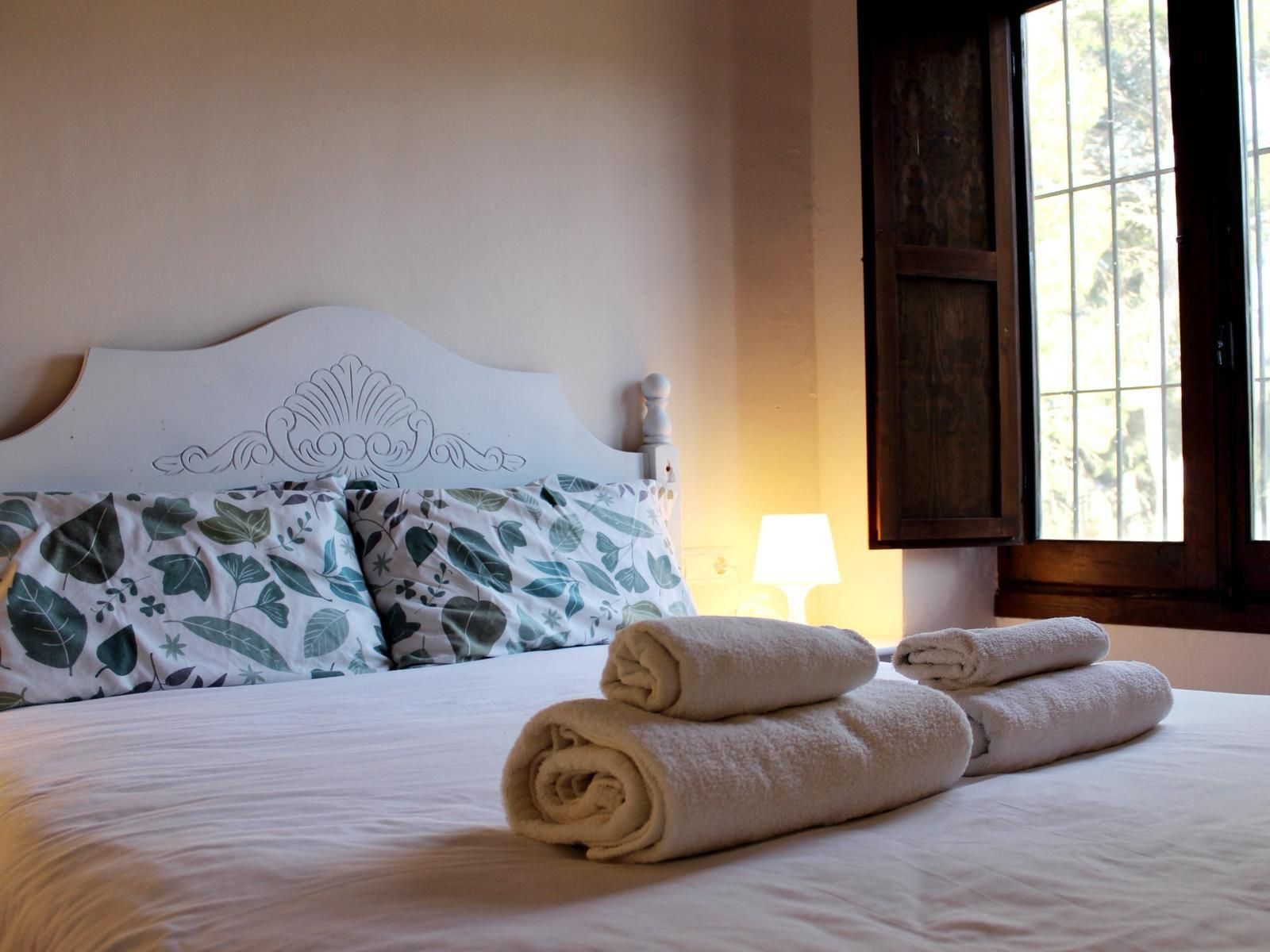 Habitación acogedora con cama blanca, toallas dobladas y luz solar que entra por una ventana abierta.