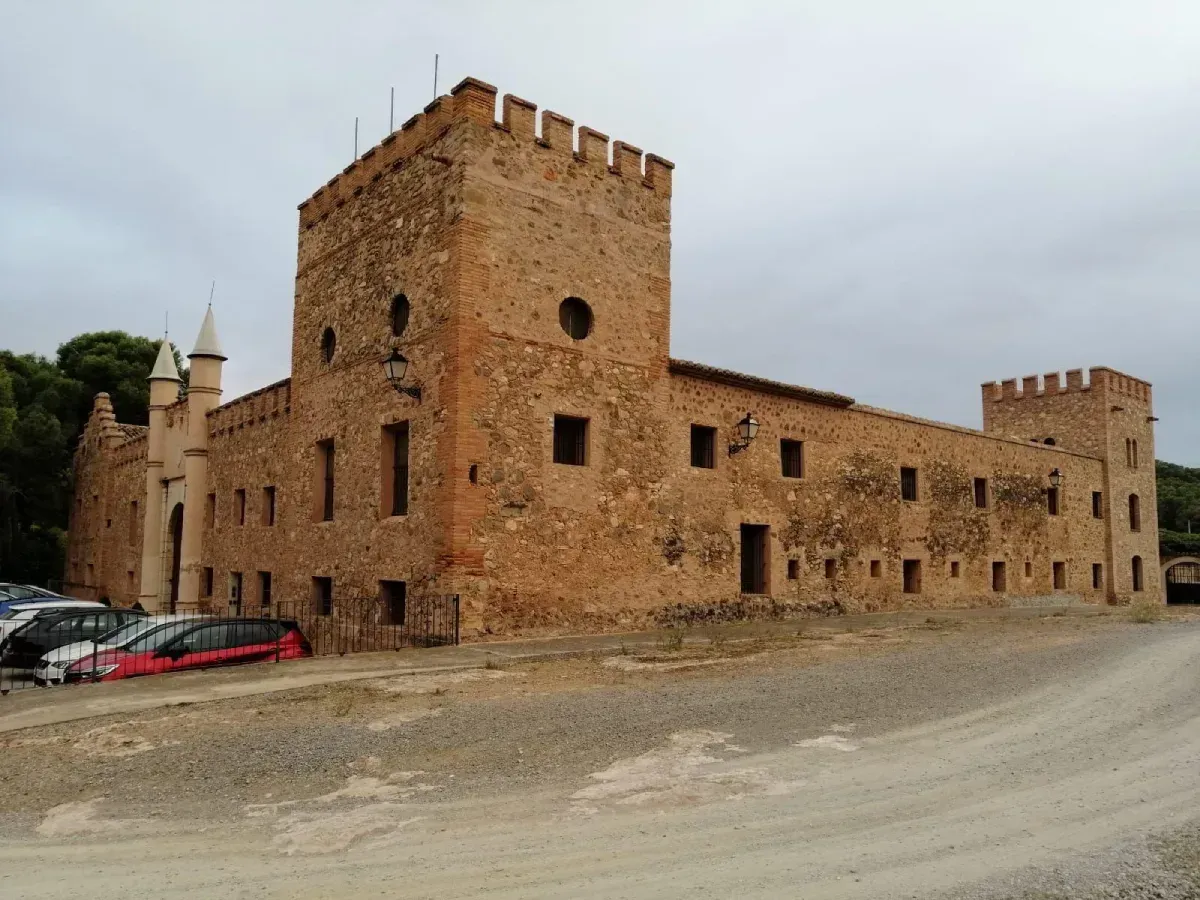 Edificio de piedra con aspecto de castillo, con almenas y coches aparcados en un terreno de grava bajo un cielo nublado.