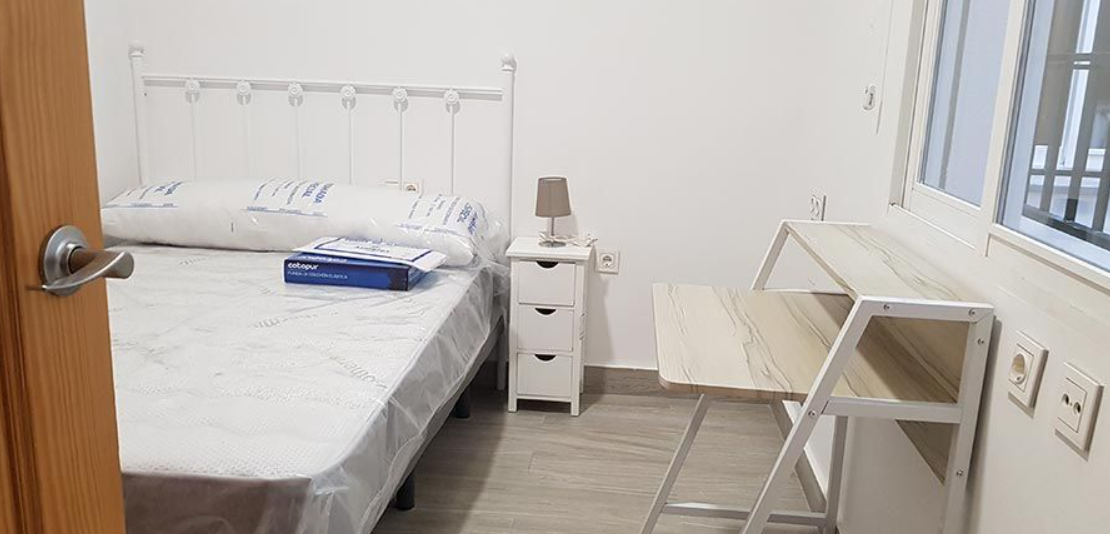 Un dormitorio con cama, mesita de noche, escritorio y ventana.