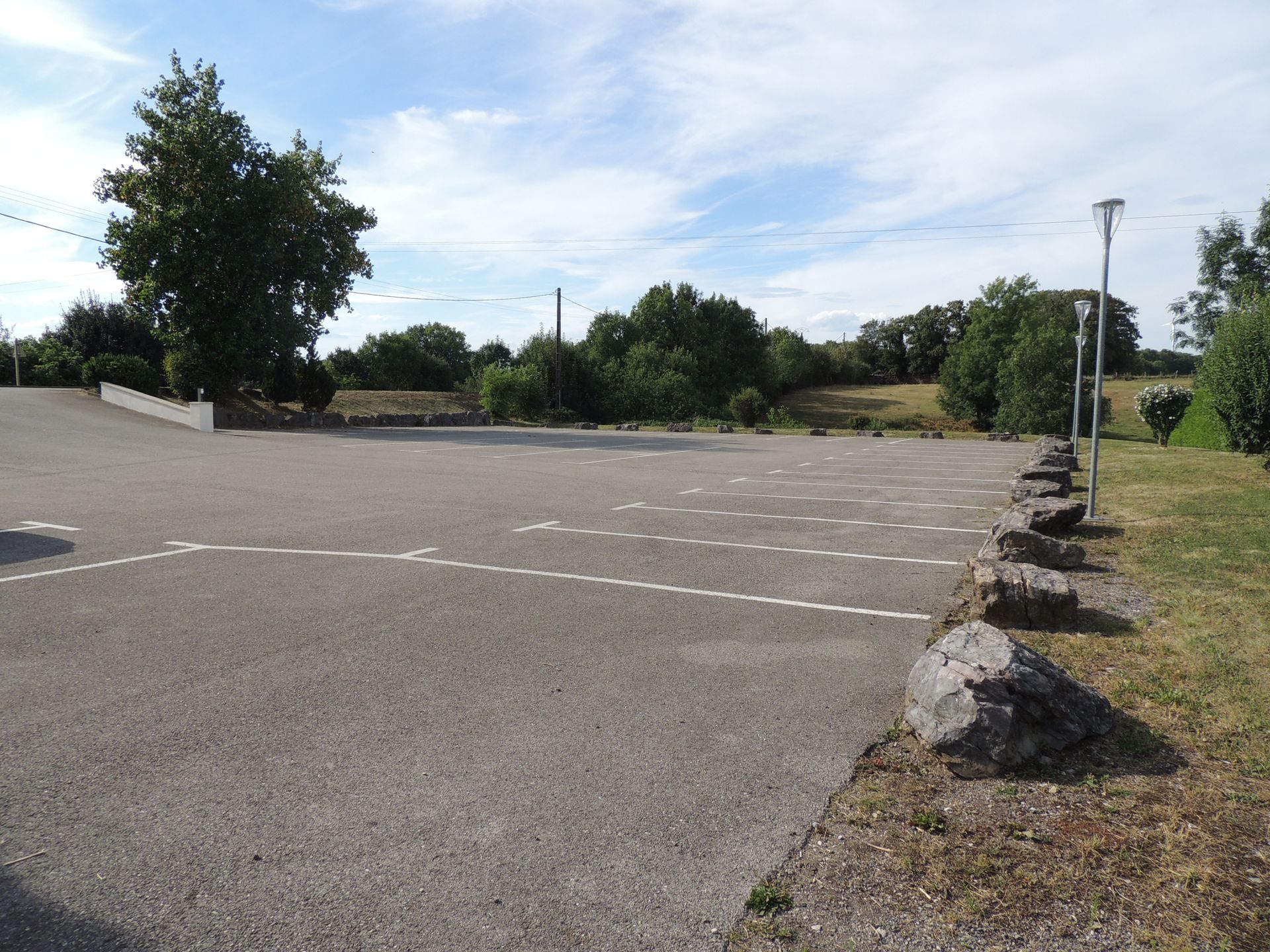 Stationnement avec places délimitées entourées d'herbe, d'arbres et d'un ciel nuageux.