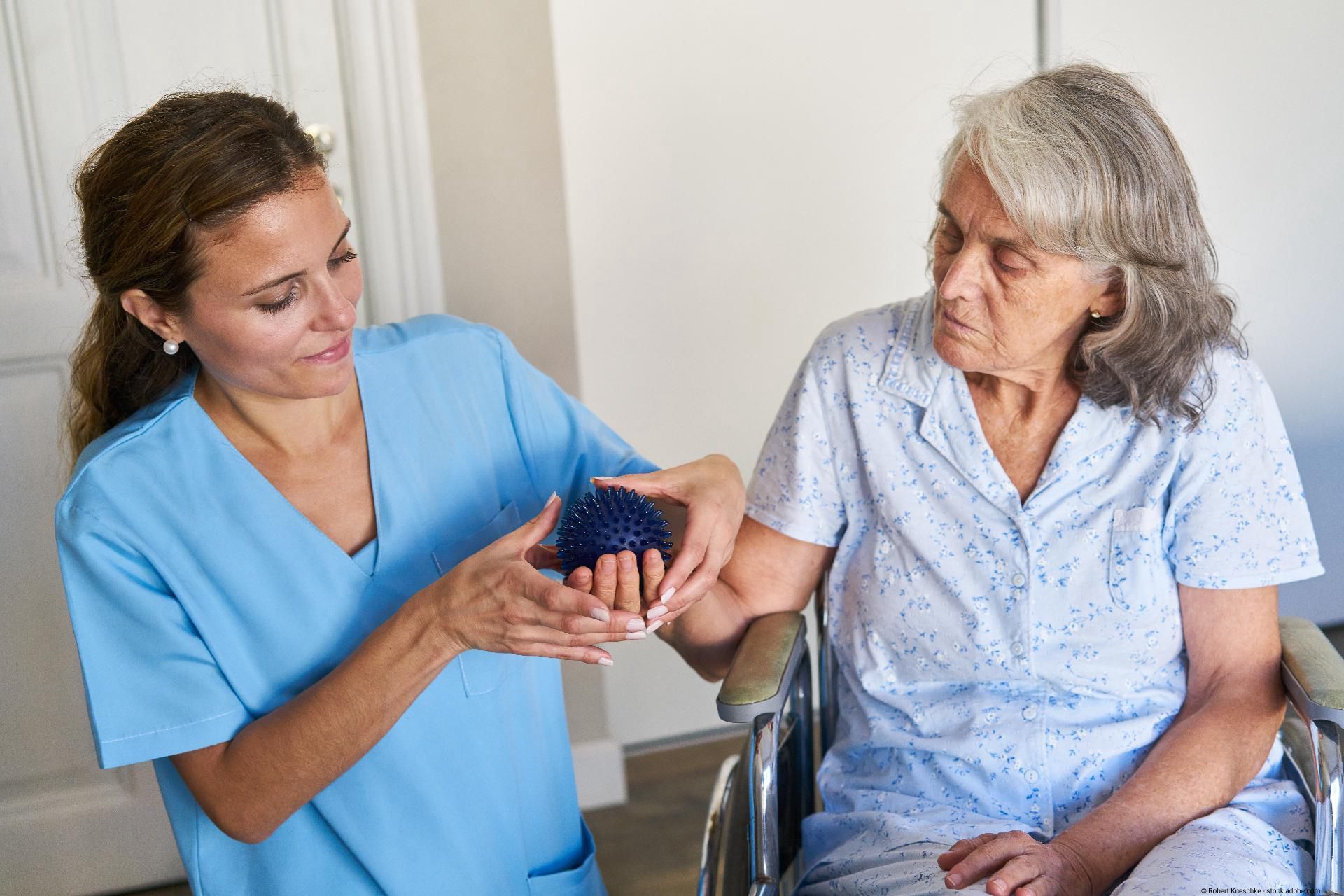 Pflegekraft erklärt einer älteren Frau die Nutzung eines Igelballs zur Handtherapie.