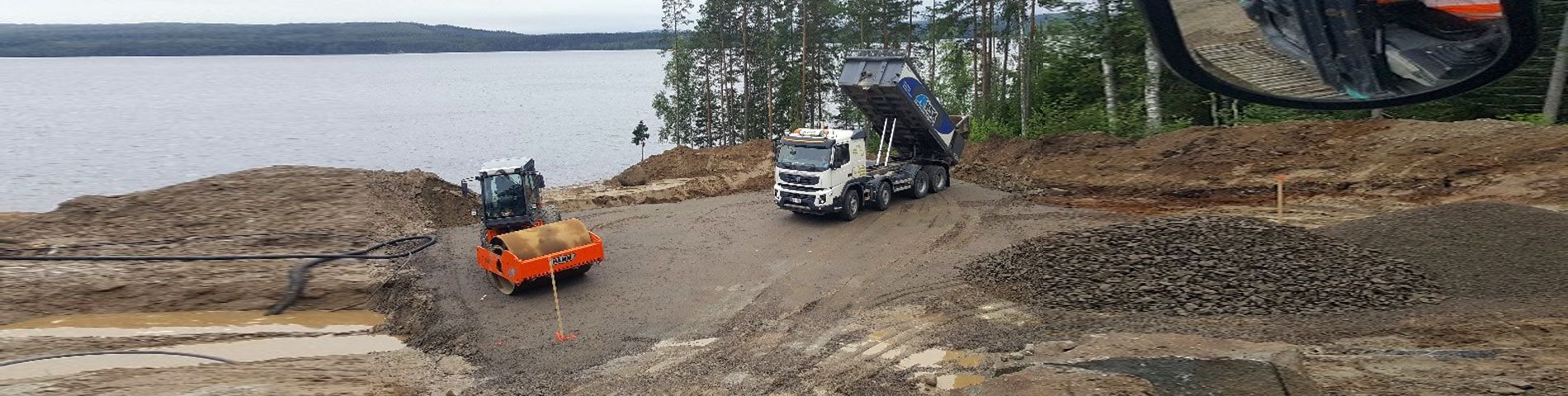 Kippiauto kippaa soraa järvenrantatyömaalla lähellä maantiivistäjää, maassa maakasojen ollessa.