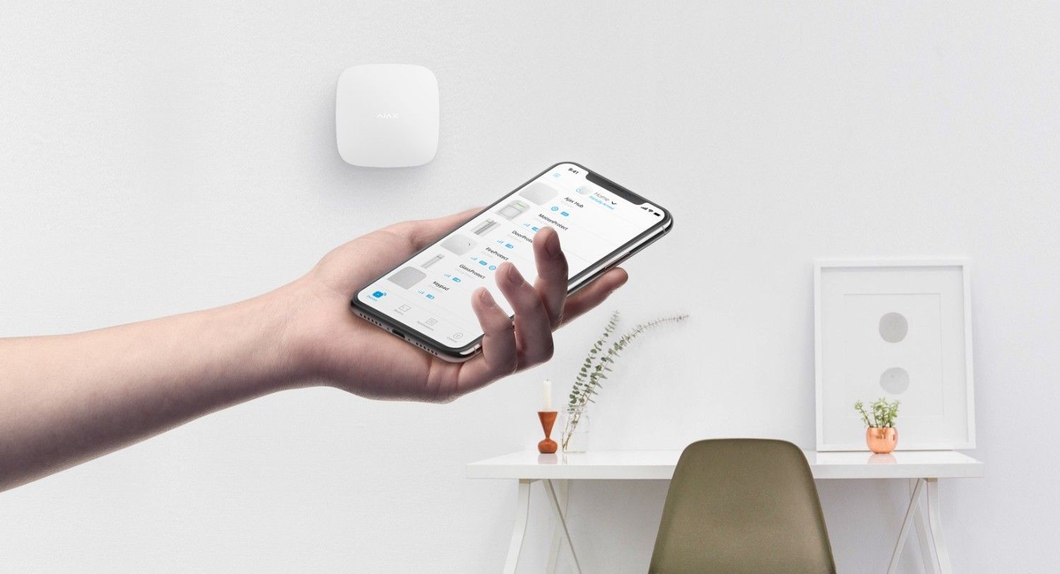 Una mano sostiene un smartphone sobre un escritorio luminoso, con una silla y unas pequeñas plantas en macetas al fondo