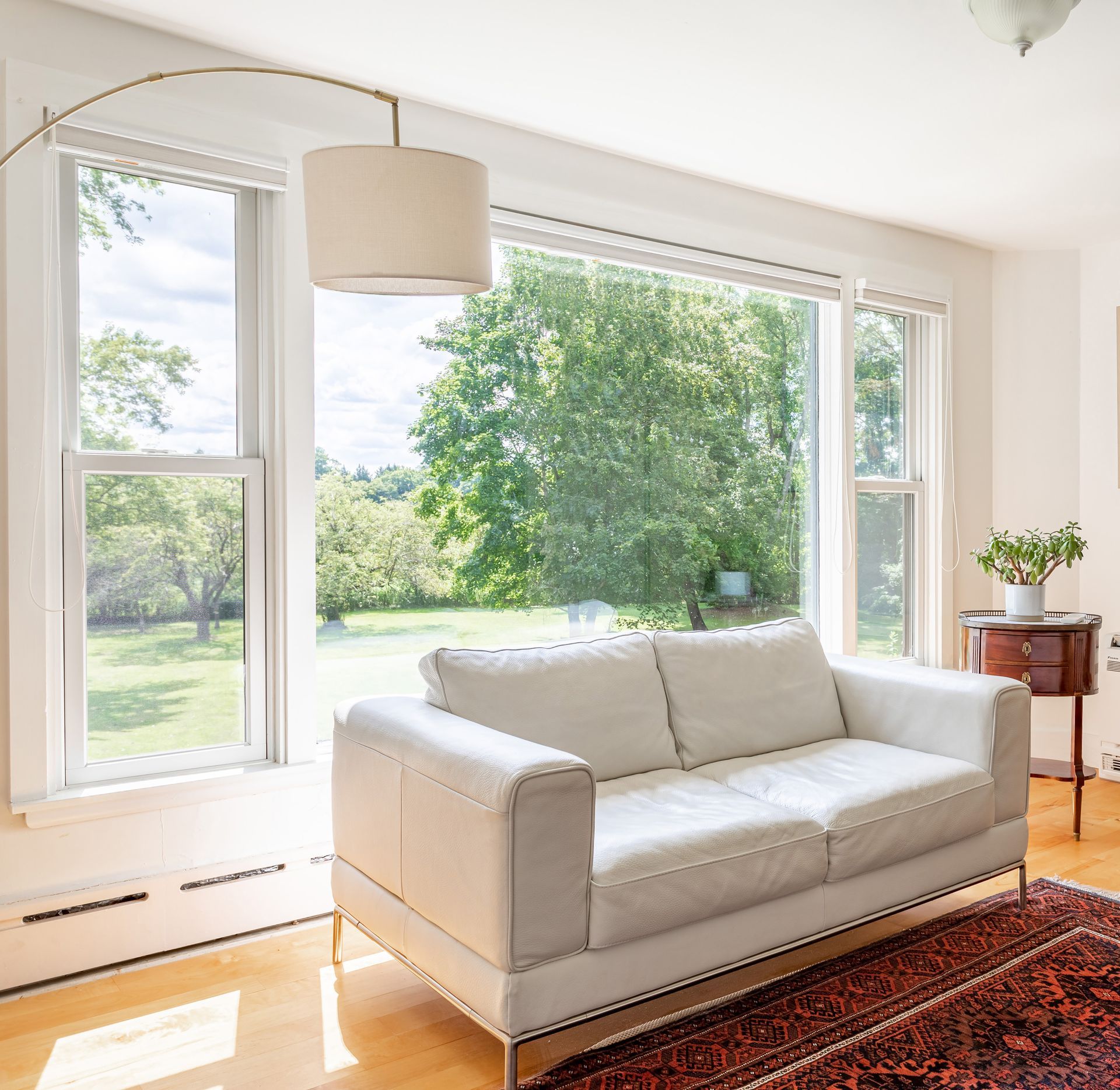 Salon lumineux avec canapé blanc, tapis rouge et grandes fenêtres donnant sur un jardin verdoyant