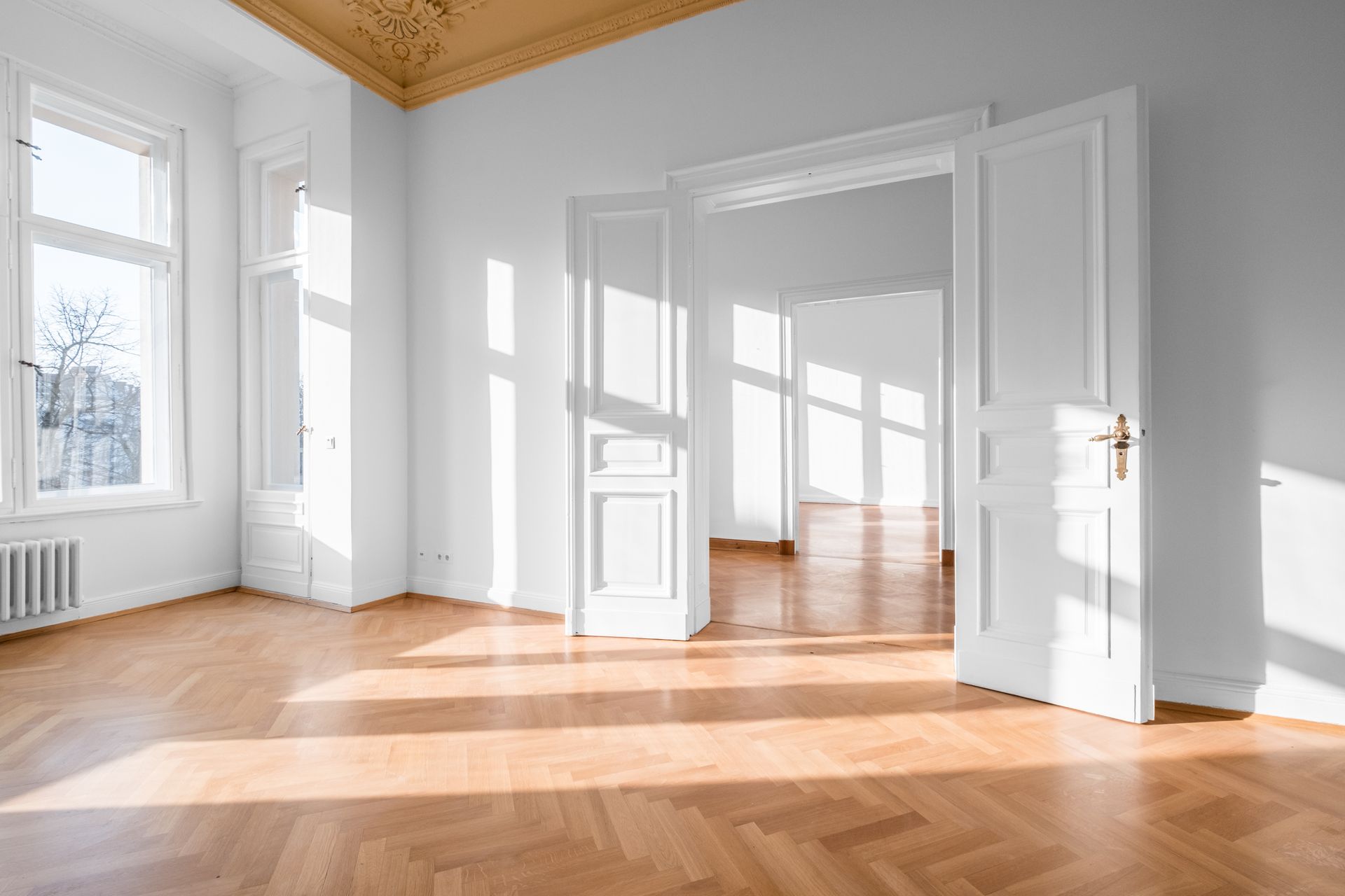 Pièce lumineuse et vide avec des murs blancs, de grandes fenêtres et un parquet baigné de soleil.