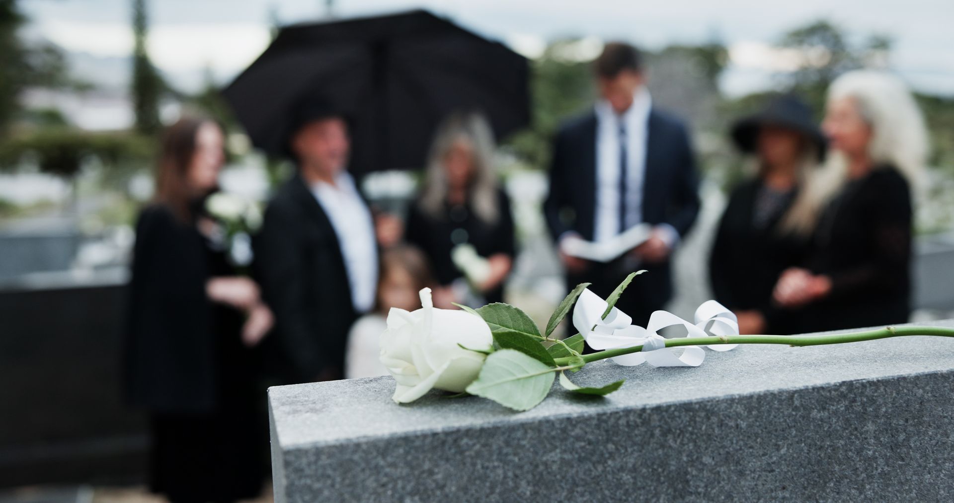 Un bouquet de roses blanches sur une pierre tombale, avec des personnes en deuil floues en arrière-plan.