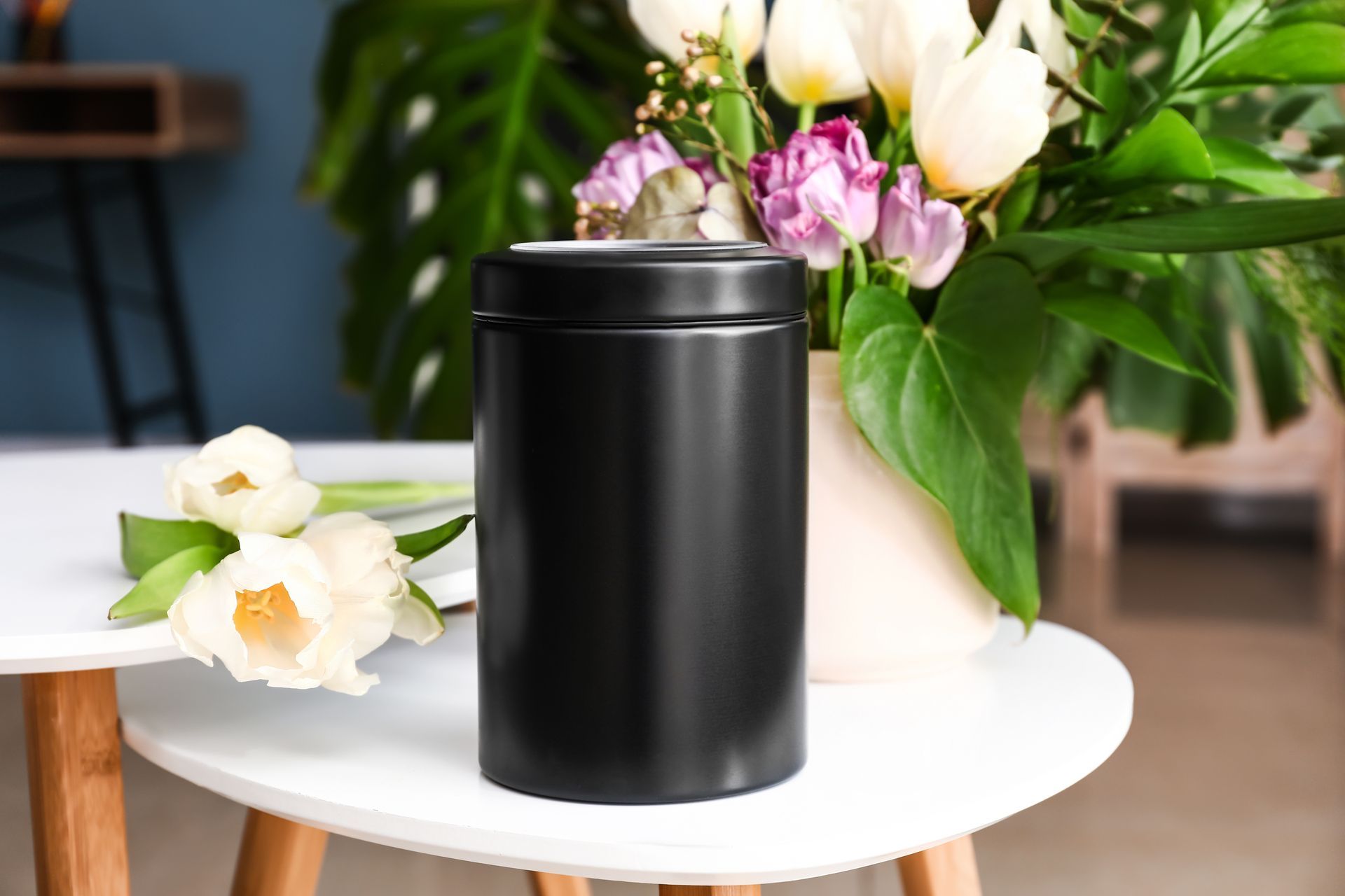 Un récipient cylindrique noir posé sur une table blanche, avec des fleurs blanches et roses en arrière-plan.