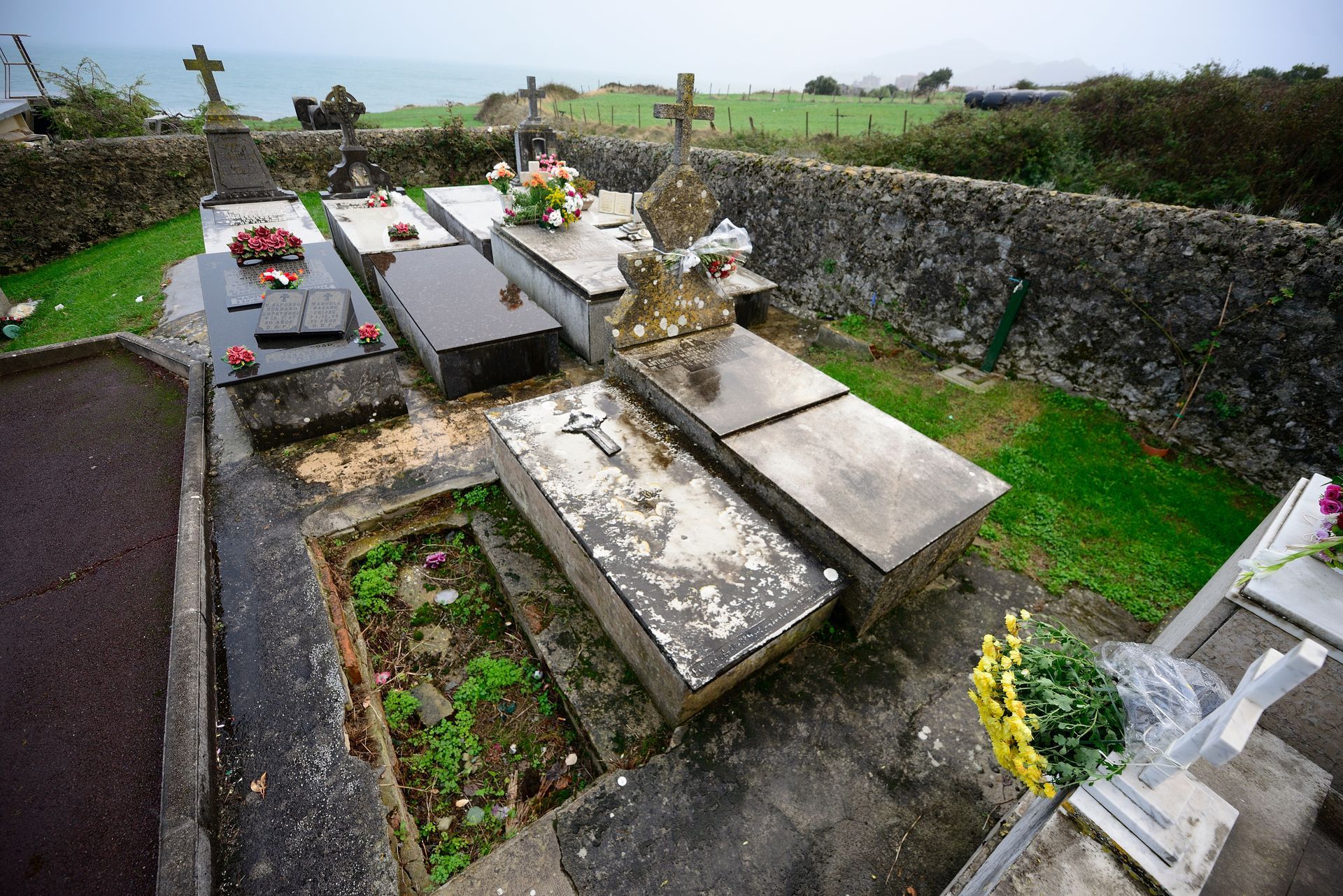 Cimetière aux tombes de pierre polie et aux fleurs, situé le long d'un mur de pierre dans un cimetière rural brumeux.