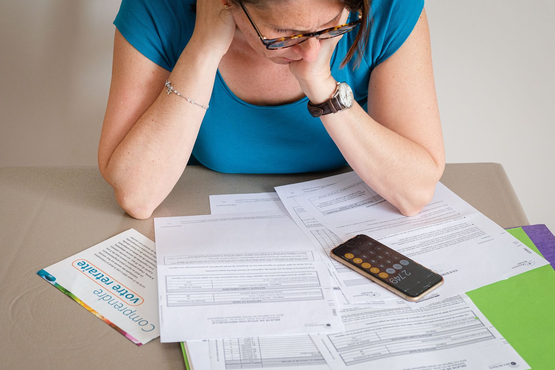 Une personne penchée sur des documents, une calculatrice posée sur une table, semble stressée.