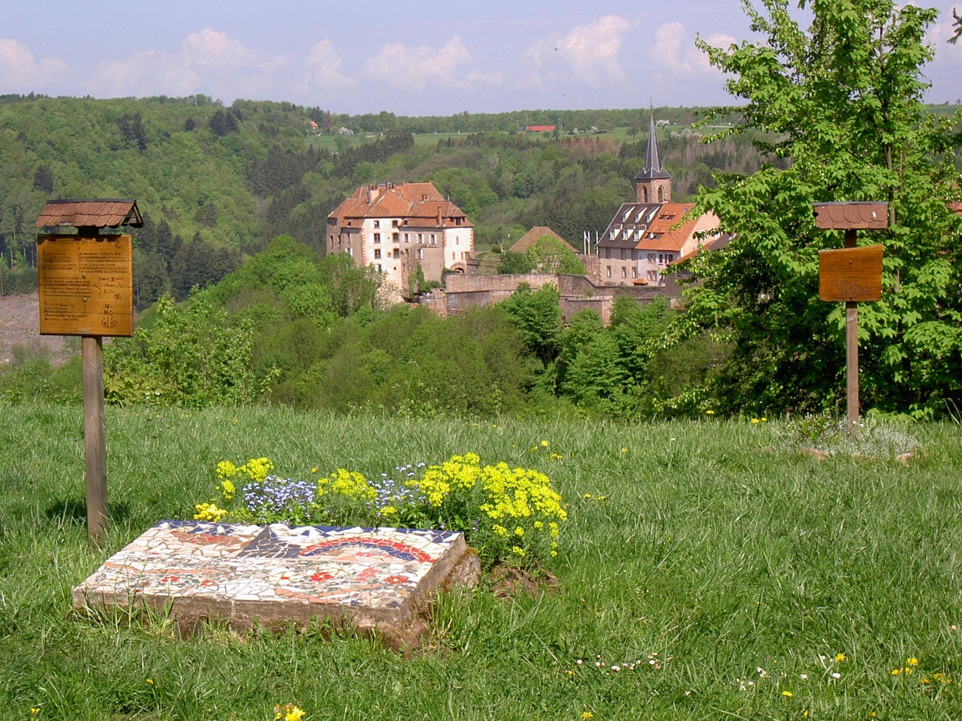 Belvédère herbeux avec panneaux, fleurs sauvages et un village au loin, sur une colline verdoyante sous un ciel partiellement nuageux.