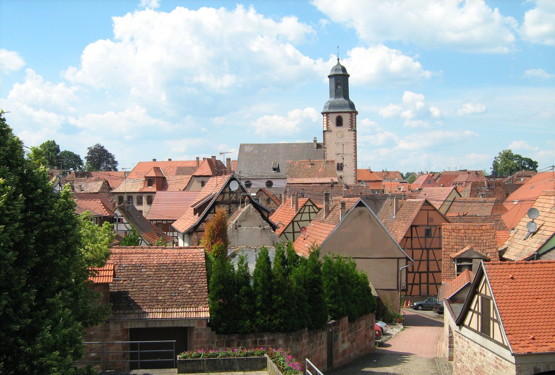 Village aux toits rouges avec un clocher en pierre sous un ciel bleu nuageux