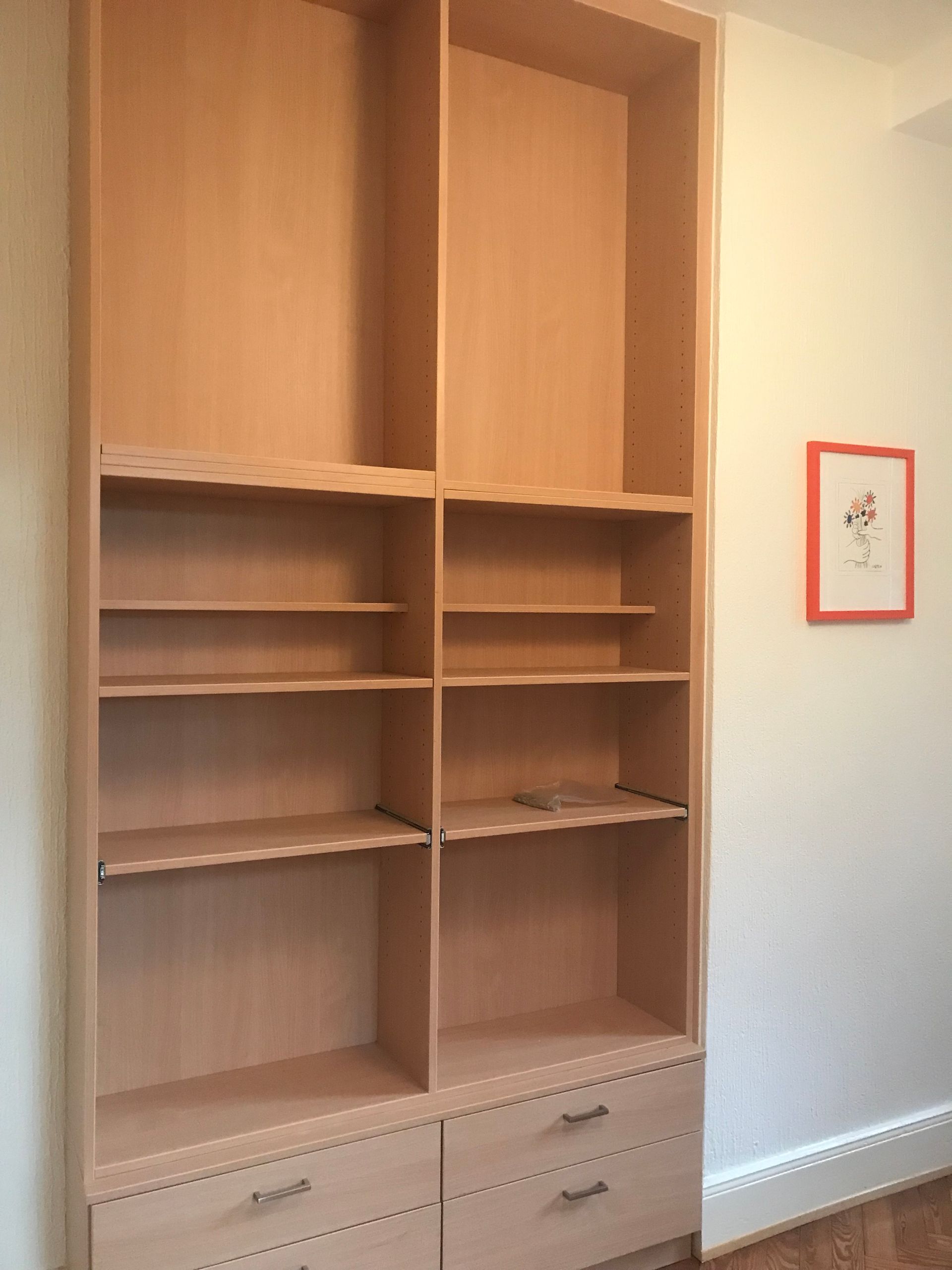 Une haute bibliothèque en bois avec tiroirs se trouve dans une pièce presque vide, à côté d'une petite estampe encadrée.