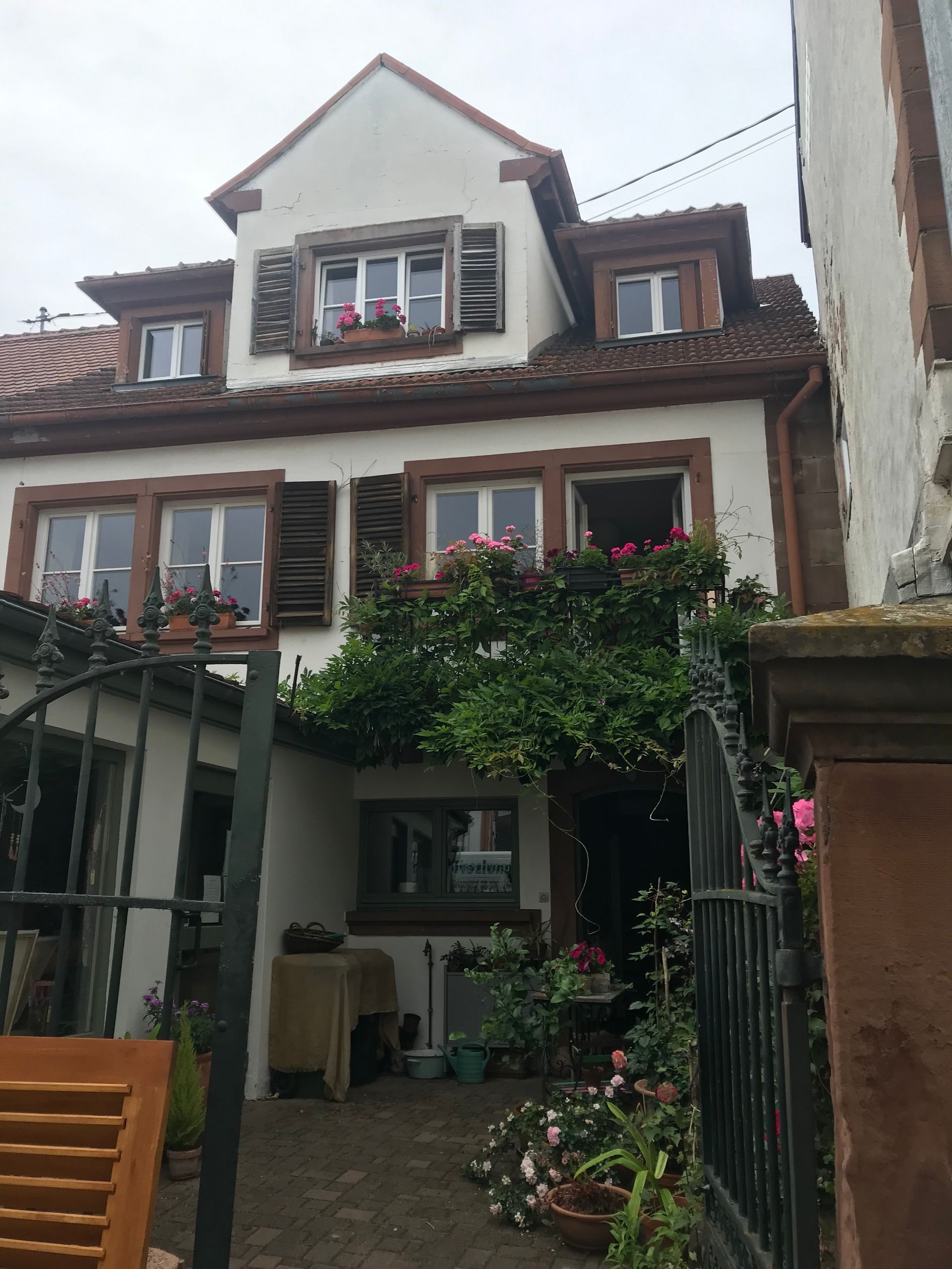 Maison blanche avec volets marron, toit de tuiles rouges, portail et jardin fleuris.