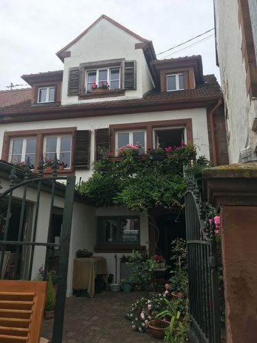 Maison blanche aux volets marron et au toit de tuiles rouges, vue à travers un portail de jardin avec des plantes devant.