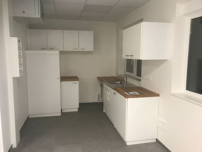 Petite cuisine vide avec placards blancs, sol gris et plans de travail en bois près d'une fenêtre.
