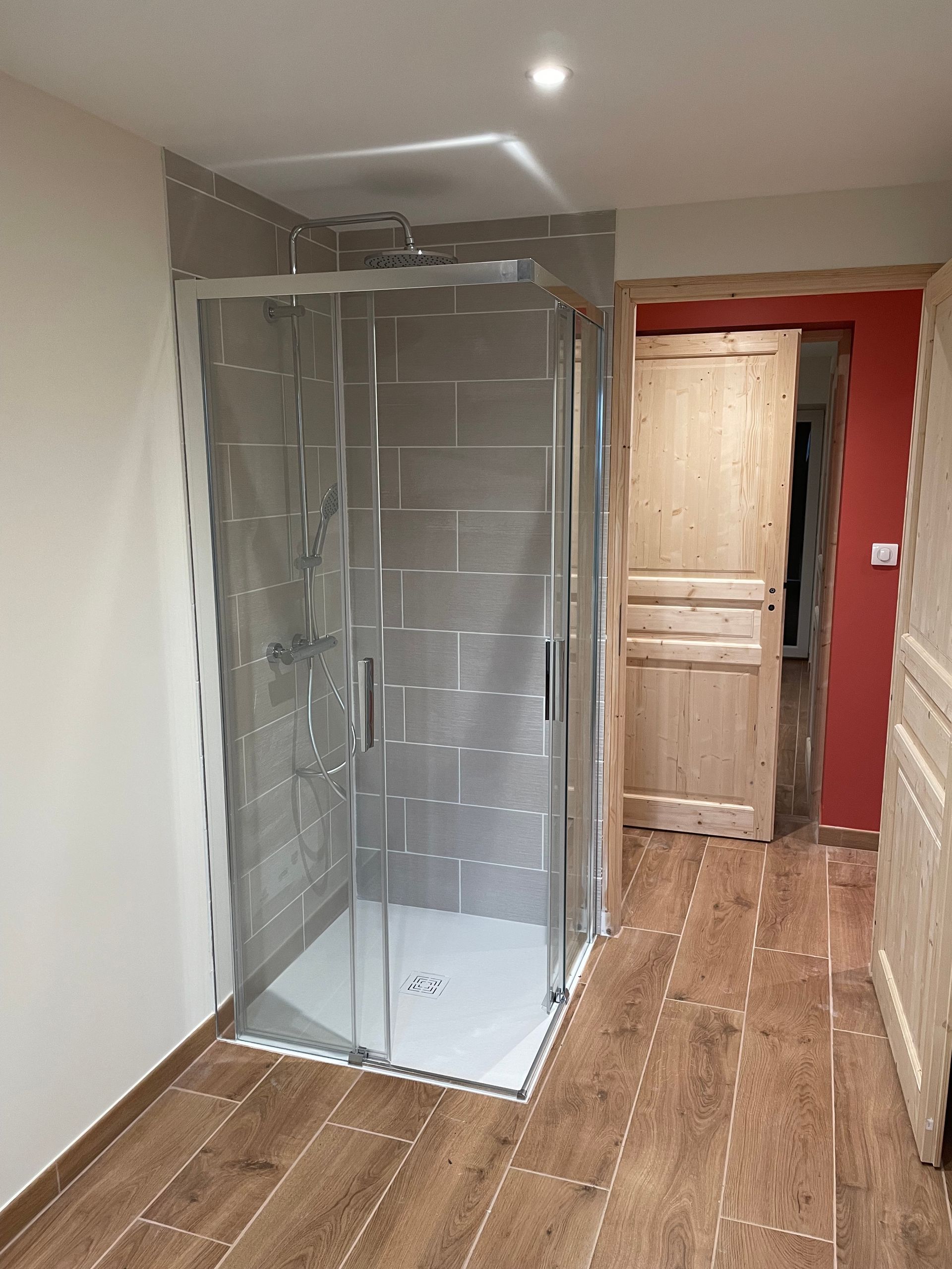 Dans une pièce au sol carrelé imitation bois et à la porte en bois, se trouve une cabine de douche vitrée aux murs carrelés de gris.