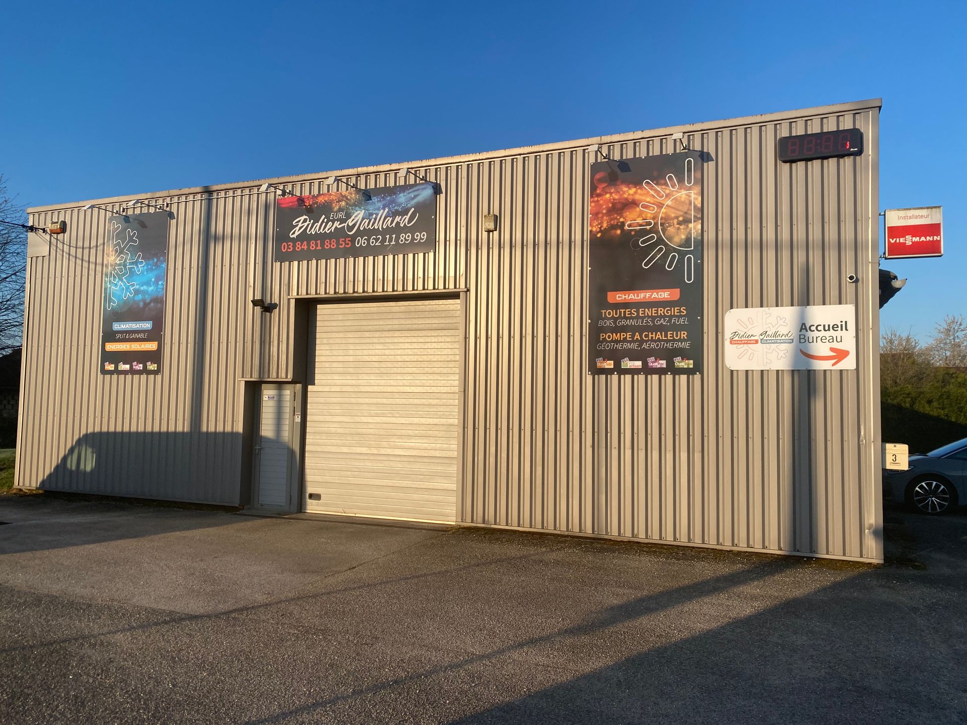 Un bâtiment commercial aux parois métalliques grises, avec une porte sectionnelle centrale et des affiches sur les murs extérieurs, sous un ciel bleu.