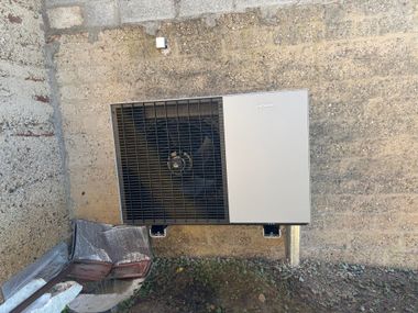 Une pompe à chaleur moderne, de couleur gris clair, montée à l'extérieur sur un mur en béton brut et texturé, près du niveau du sol.