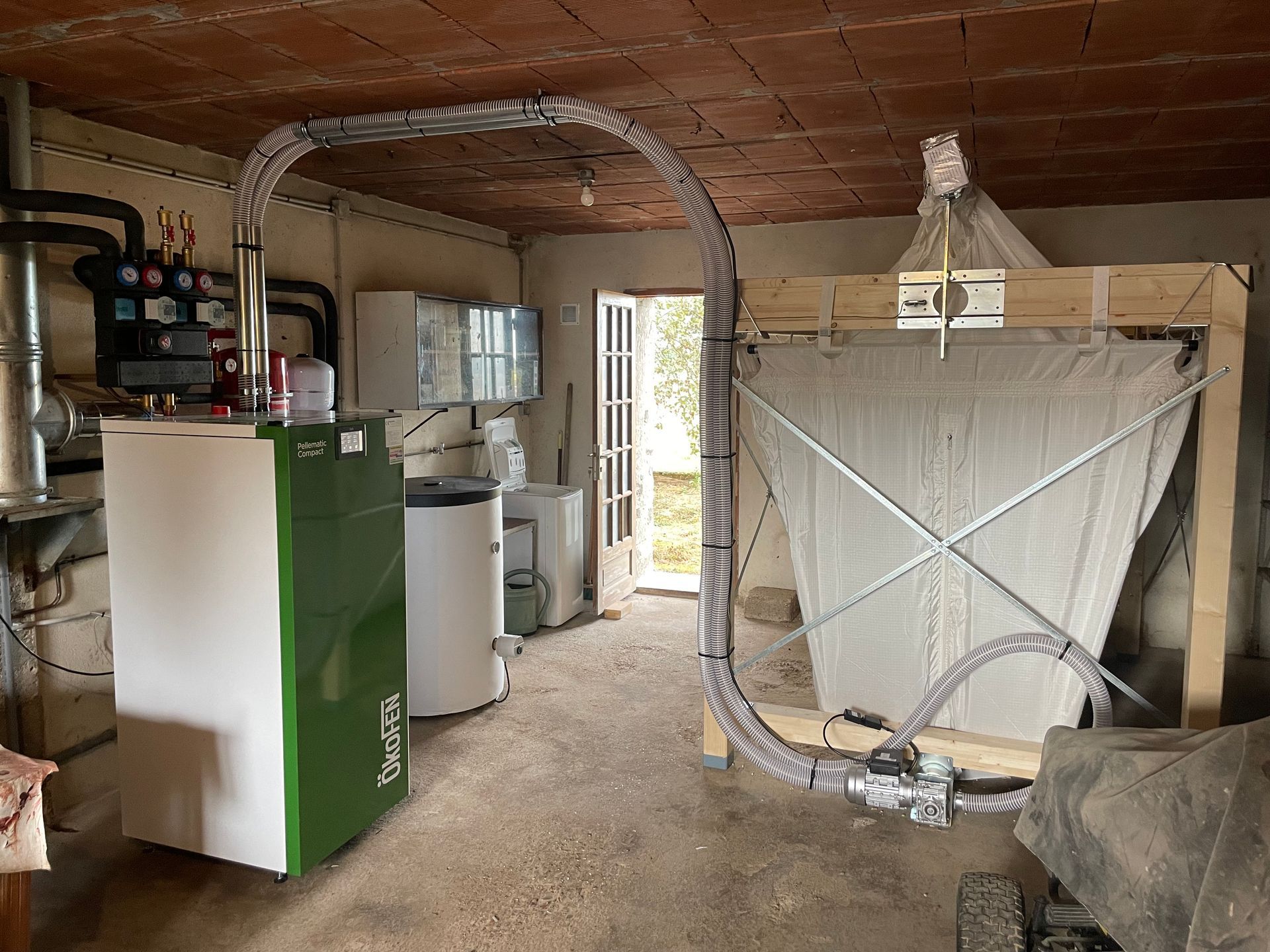 Une chaudière à granulés verts est reliée à un silo de stockage en tissu par un tuyau flexible dans une buanderie au plafond en briques.
