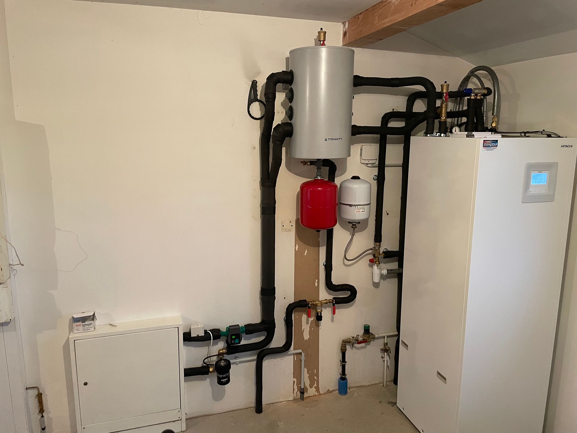 Un système de pompe à chaleur avec une unité de plancher blanche, un réservoir tampon gris et un vase d'expansion rouge monté sur un mur blanc.