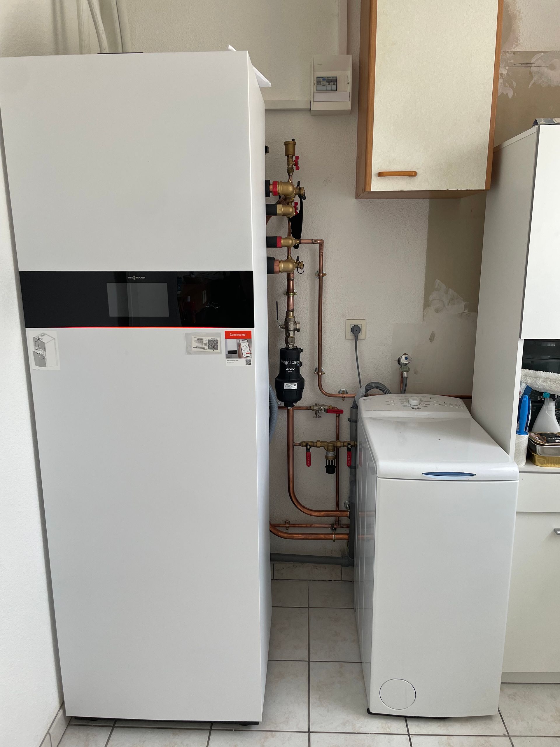 Dans une buanderie, une grande pompe à chaleur blanche se dresse à côté d'une machine à laver, avec des tuyaux en cuivre fixés au mur.