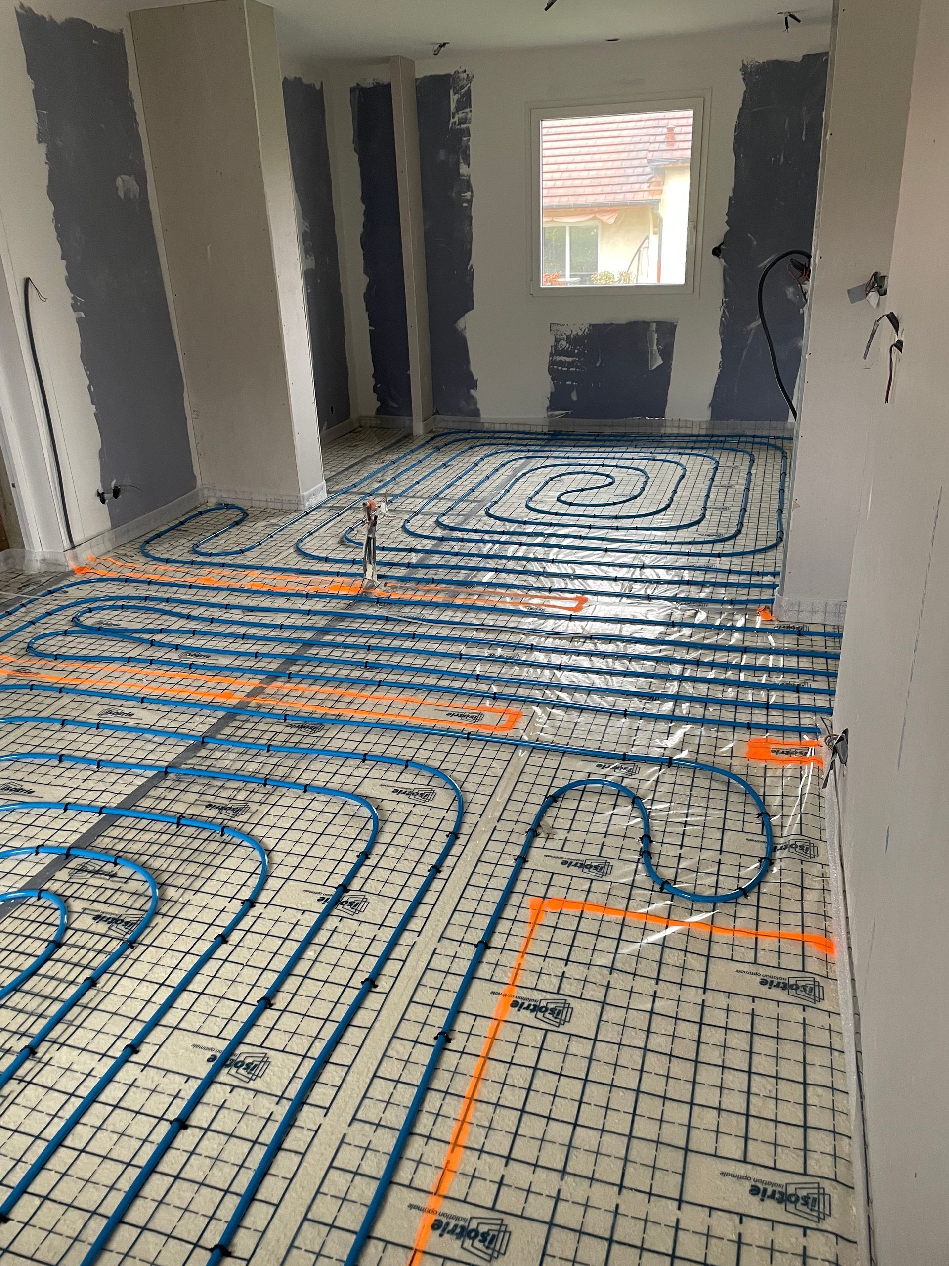 Des tuyaux de chauffage au sol, hydroniques bleus sont disposés en boucles sur une sous-couche à motif quadrillé dans une pièce en construction.