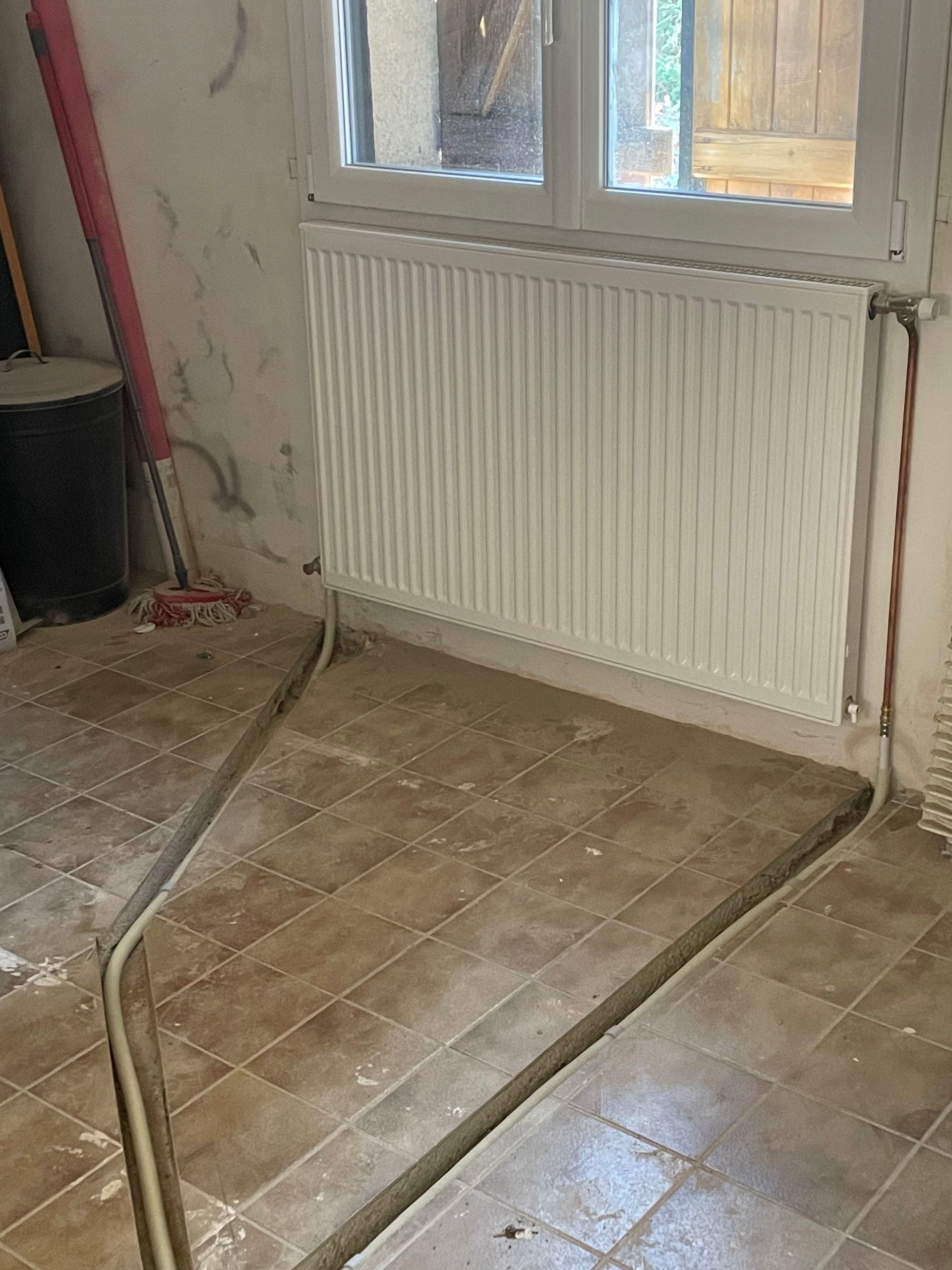 Un radiateur blanc fixé sous une fenêtre dans une pièce au sol carrelé et aux tuyaux apparents.